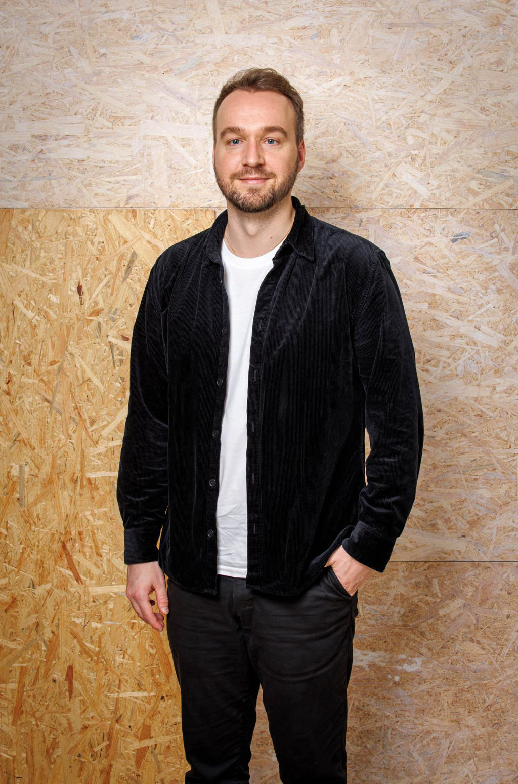 En man med kort hår och skägg står mot en vägg av träpanel, har svart jacka, vit t-shirt, svarta byxor och ena handen i fickan.