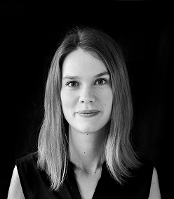 En kvinna med rakt axellångt hår och neutralt ansiktsuttryck ser in i kameran mot en svart bakgrund.