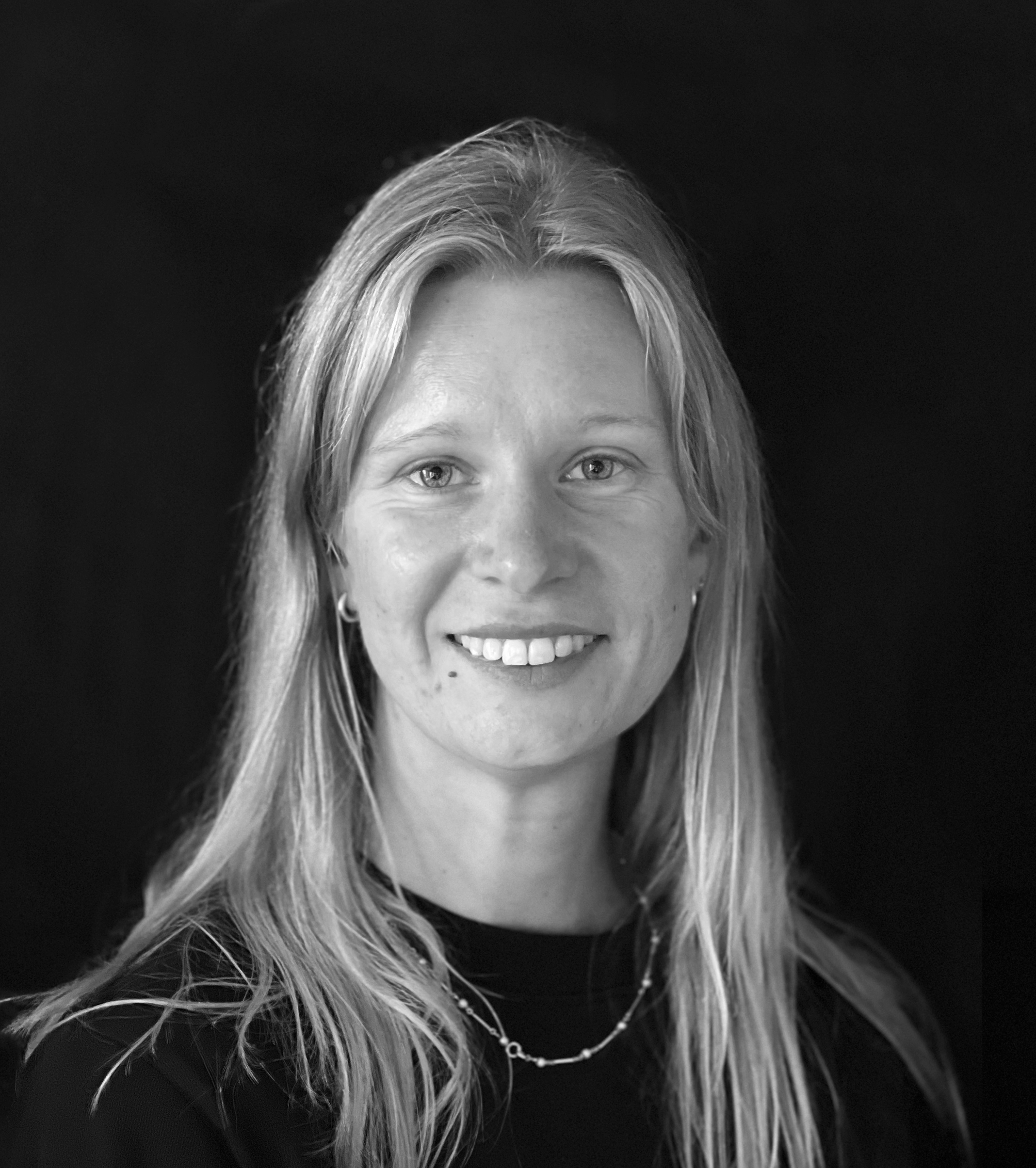En kvinna med långt ljust hår, mörk överdel och halsband ler mot kameran framför en mörk bakgrund.