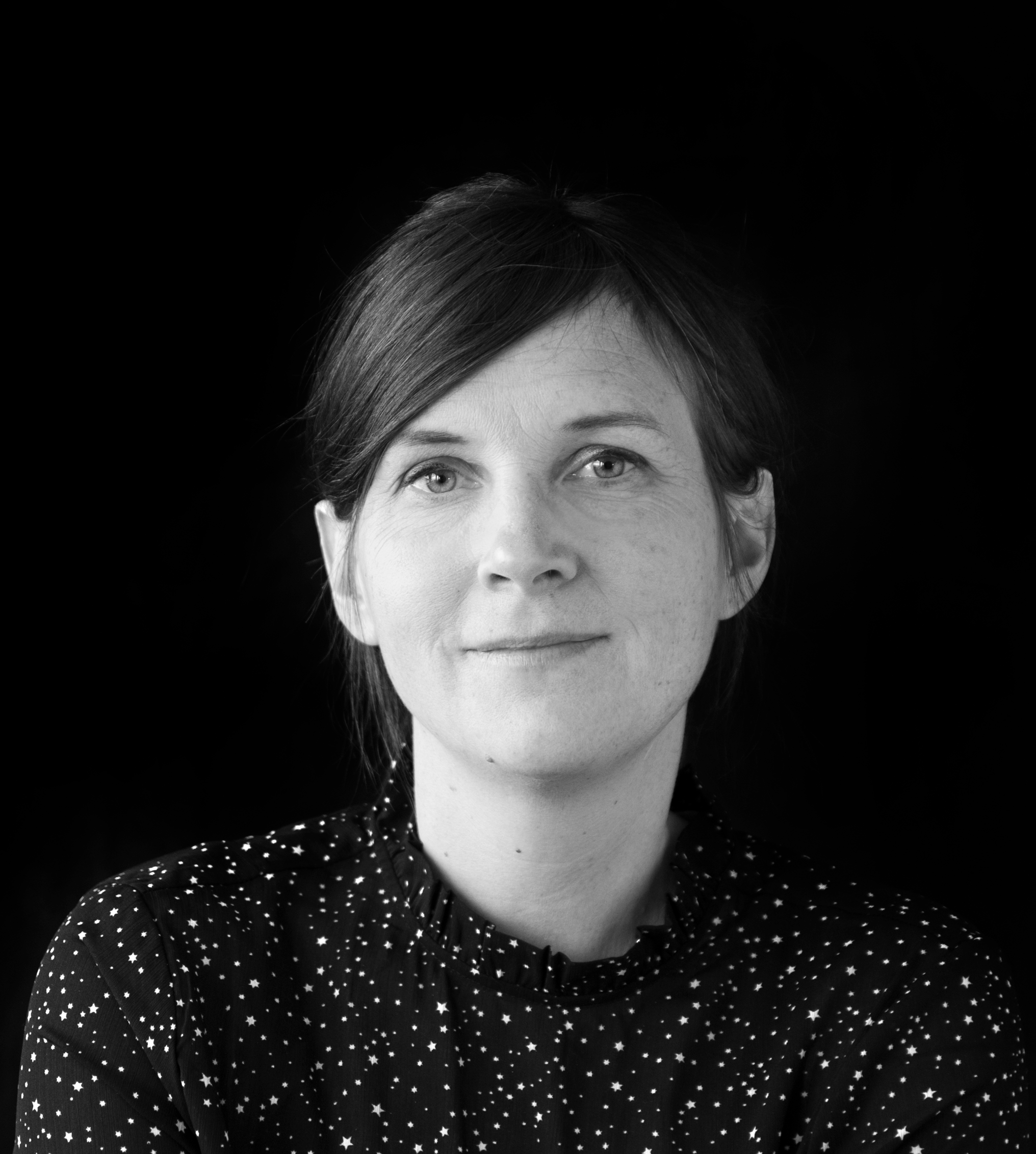 En kvinna med rakt brunt hår och neutral min har på sig en mörk skjorta med vitt stjärnmönster mot svart bakgrund.