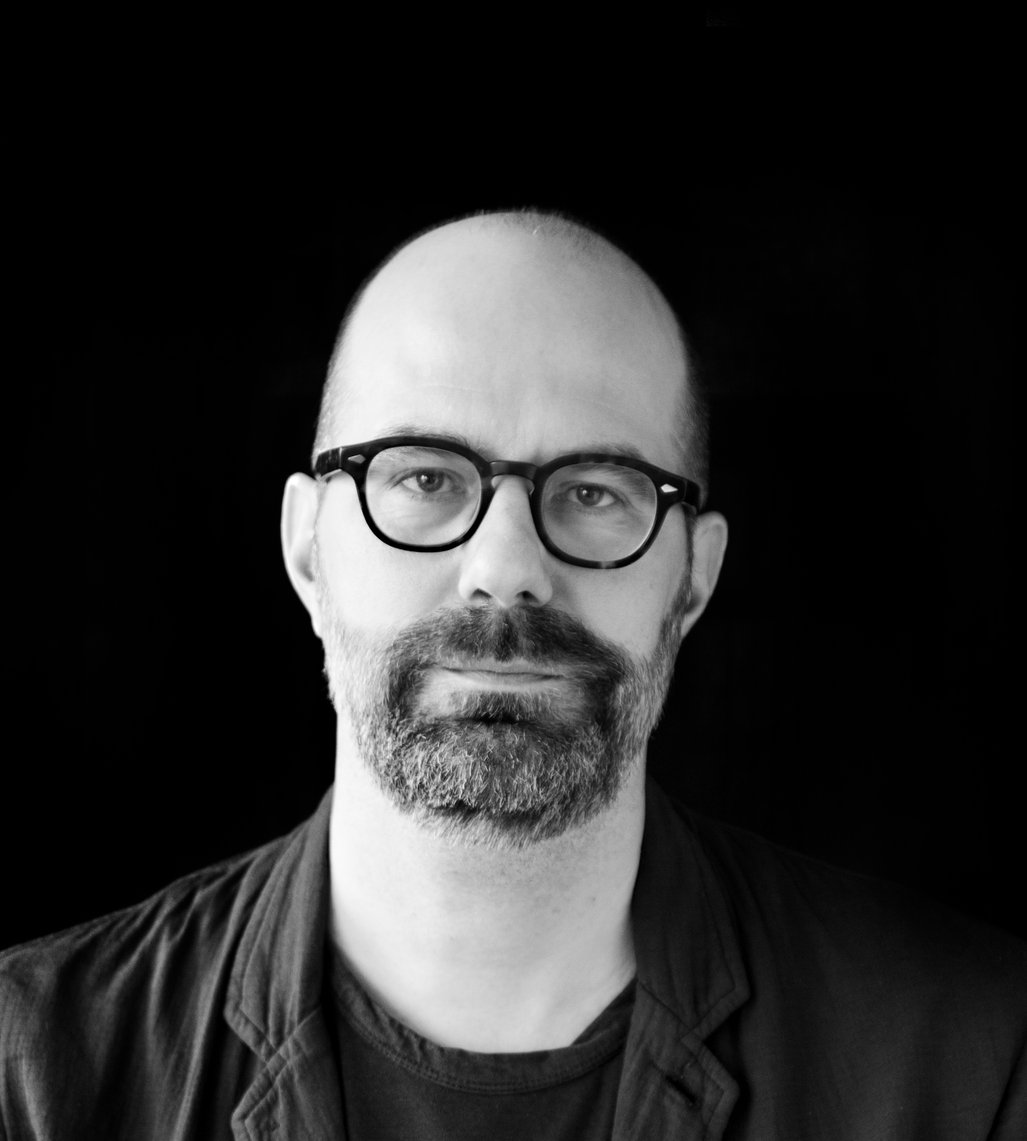 En man med skägg och glasögon har på sig en mörk jacka och skjorta, tittar rakt mot kameran mot en svart bakgrund.