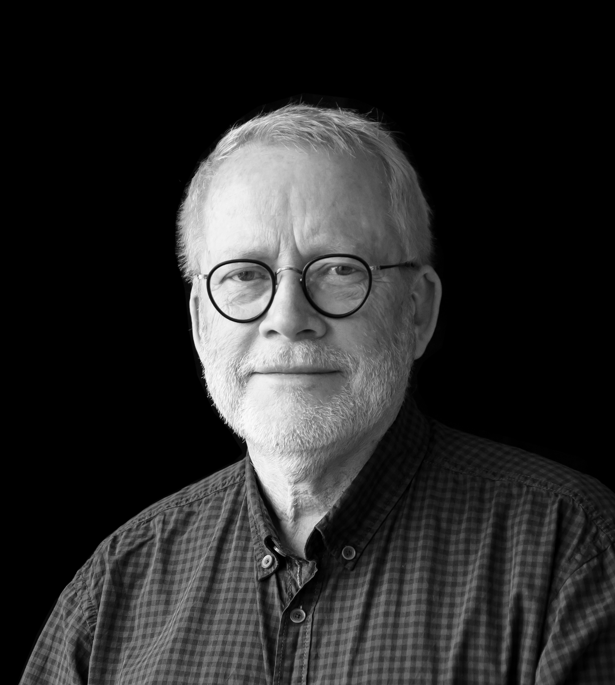 En äldre man med glasögon och skägg bär en rutig skjorta och står mot en svart bakgrund.