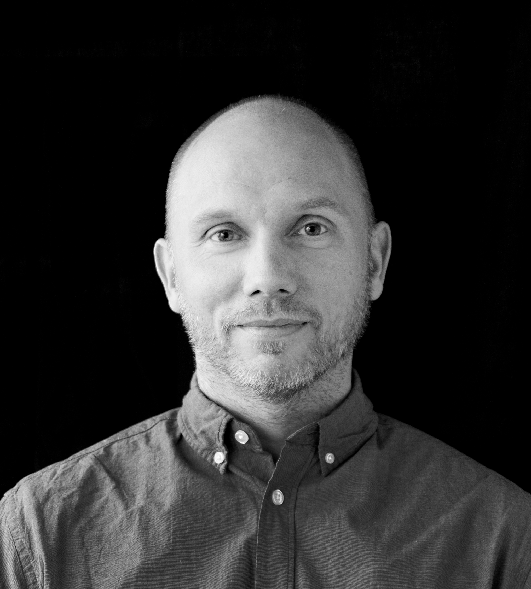 En man med rakat huvud och kort skägg, iklädd en skjorta, ser mot kameran framför en svart bakgrund.