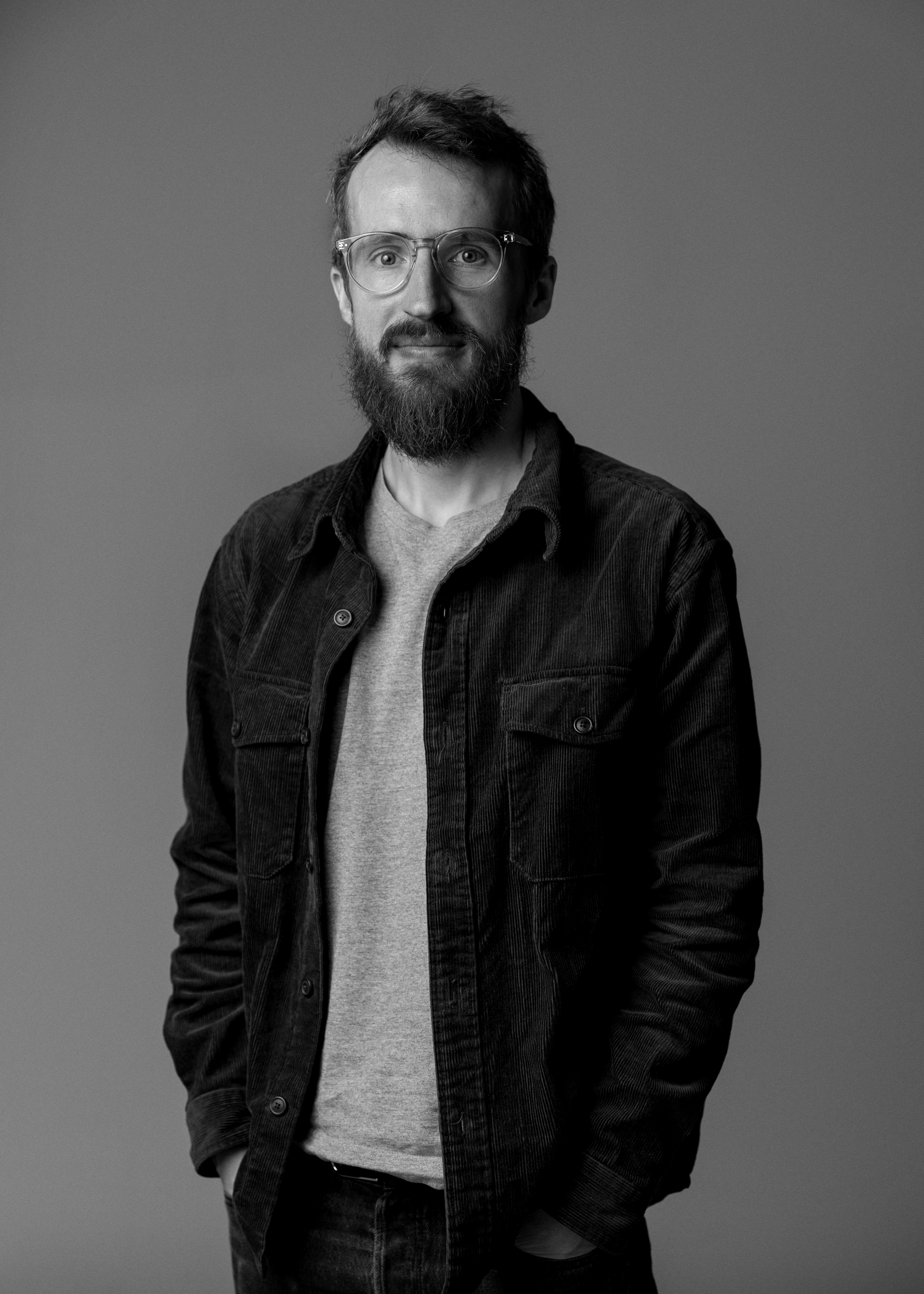 En man med glasögon och skägg står mot en enkel bakgrund, har en skjorta över en t-shirt och ena handen i fickan.