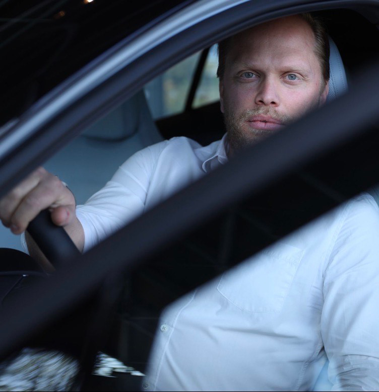 En man med skägg och vit skjorta sitter i förarsätet på en bil och tittar ut genom det öppna fönstret.