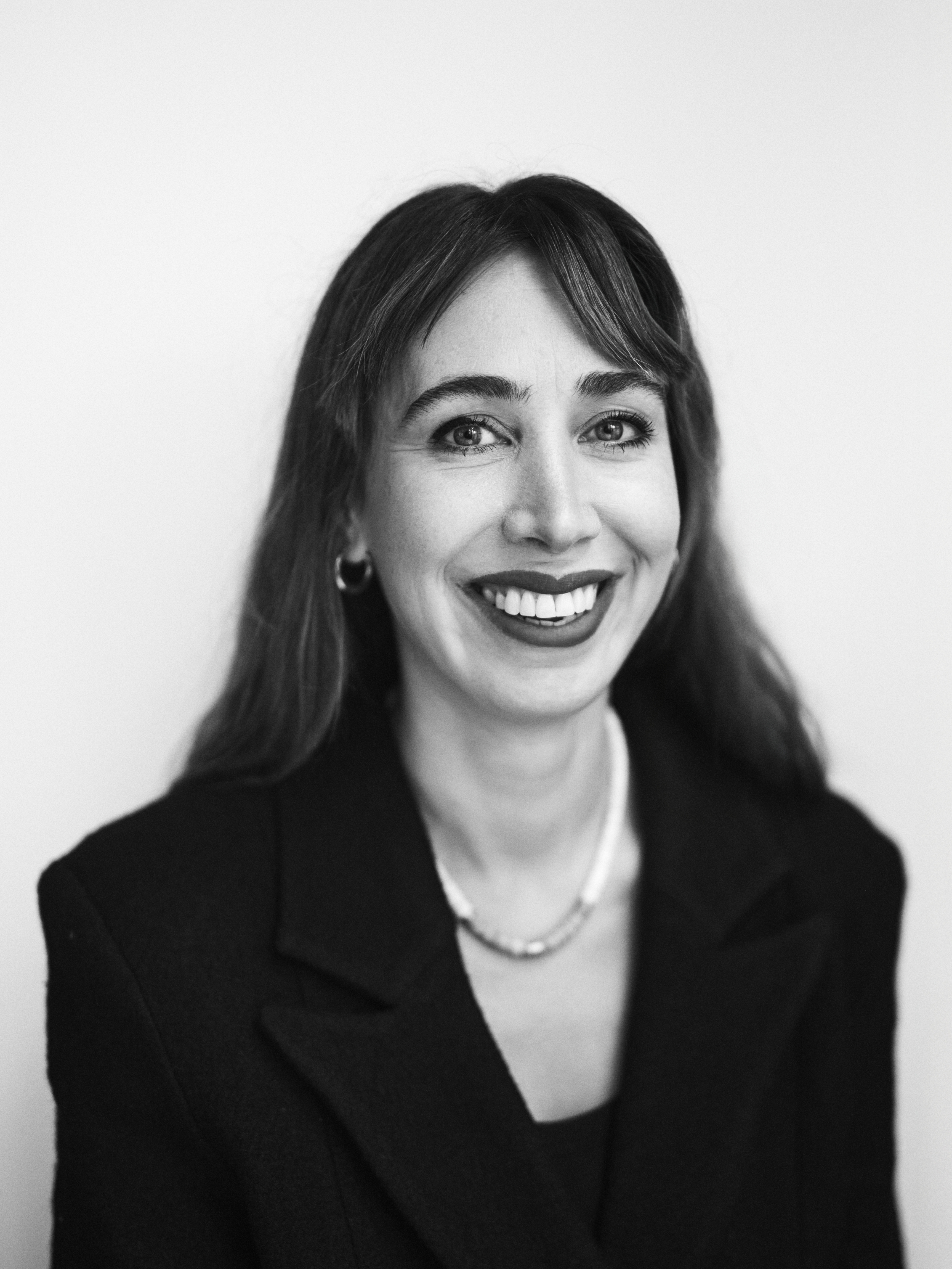 Porträtt i svartvitt av en kvinna med långt hår, kavaj, stora örhängen och halsband som ler mot kameran, enkel bakgrund.