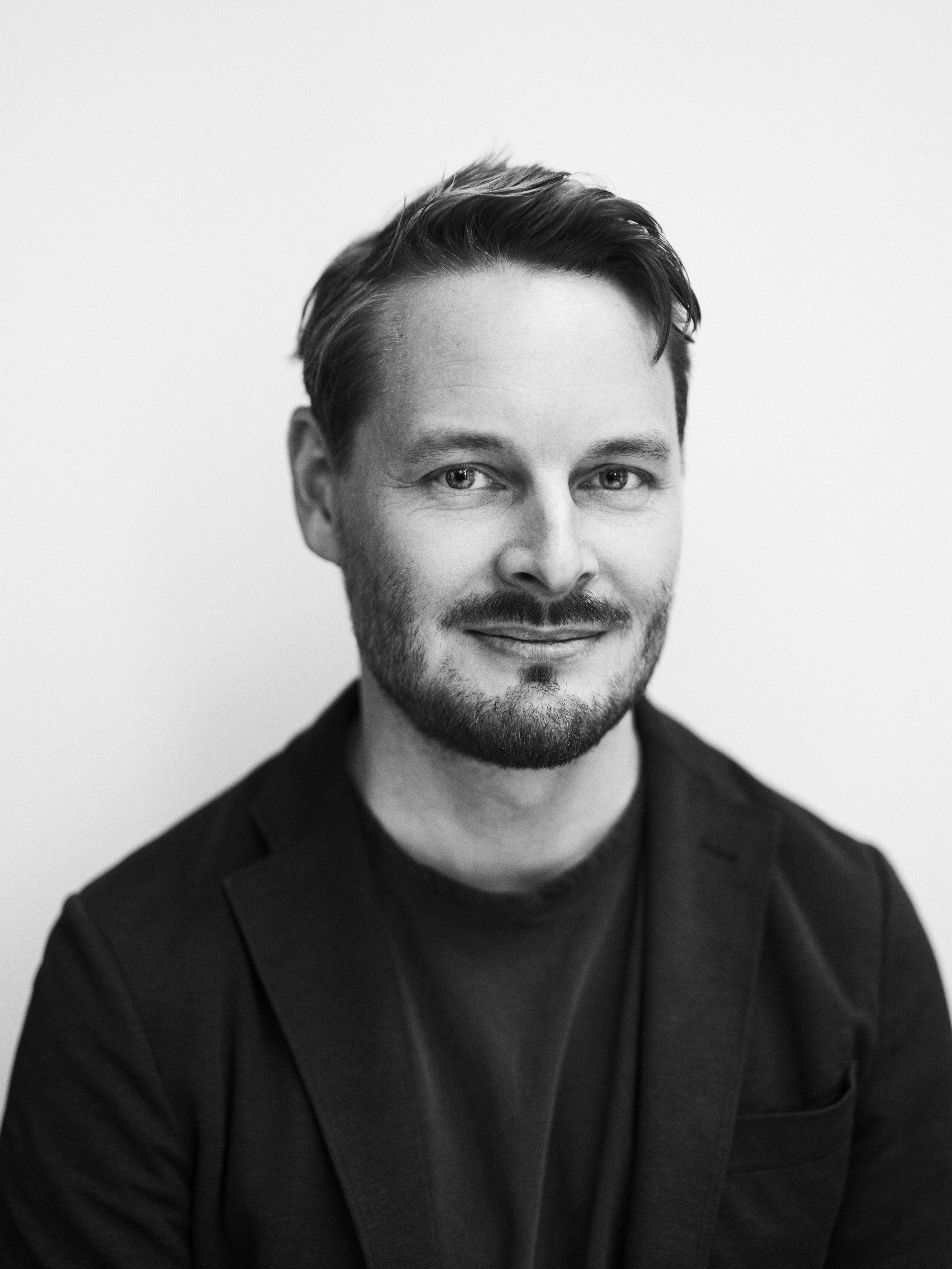 En man med kort hår och skägg har på sig en mörk jacka och skjorta, ler svagt och ser mot kameran framför en enkel bakgrund.