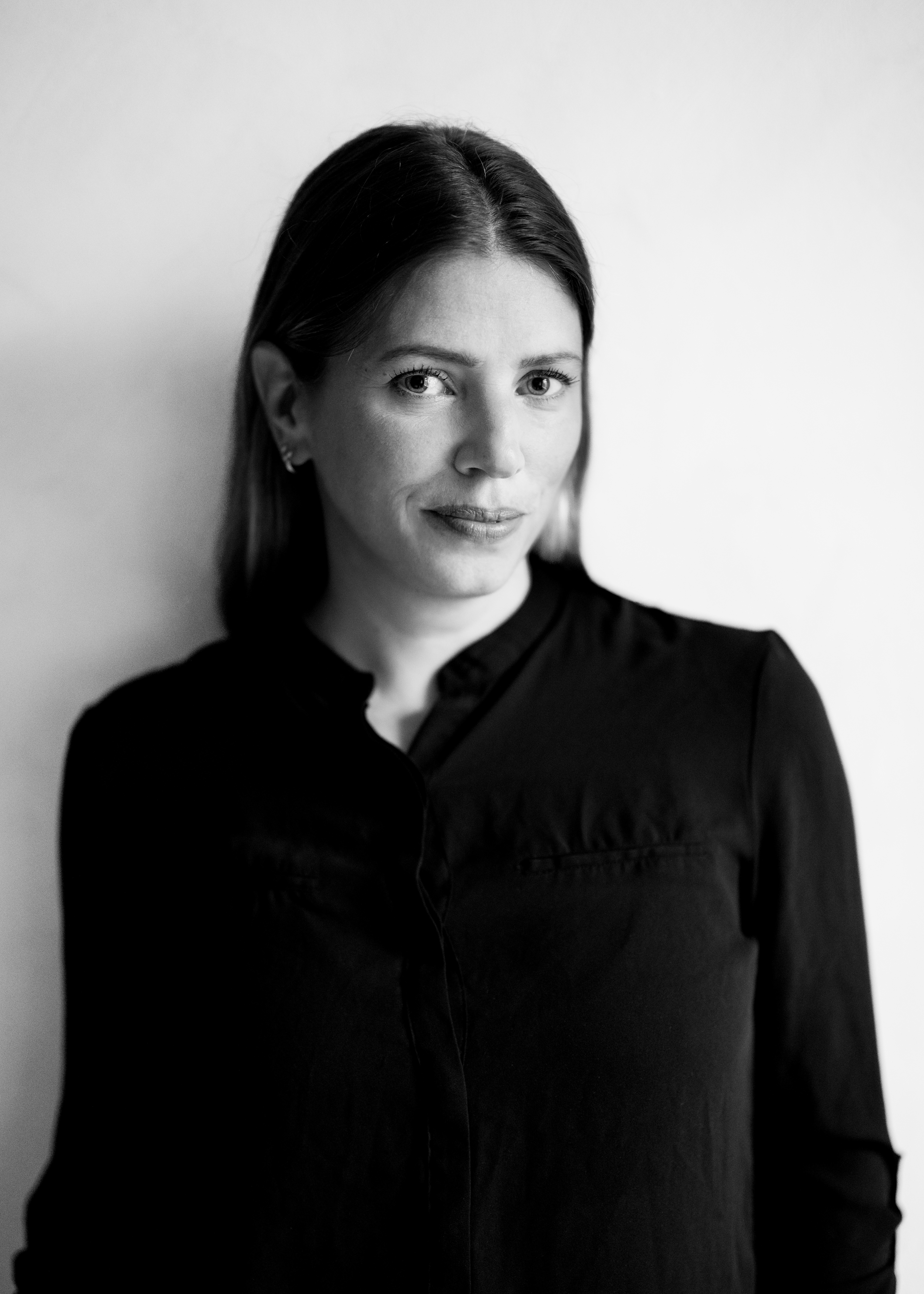 En kvinna med rakt hår och mörk skjorta står mot en ljus bakgrund och tittar in i kameran. Bilden är i svartvitt.