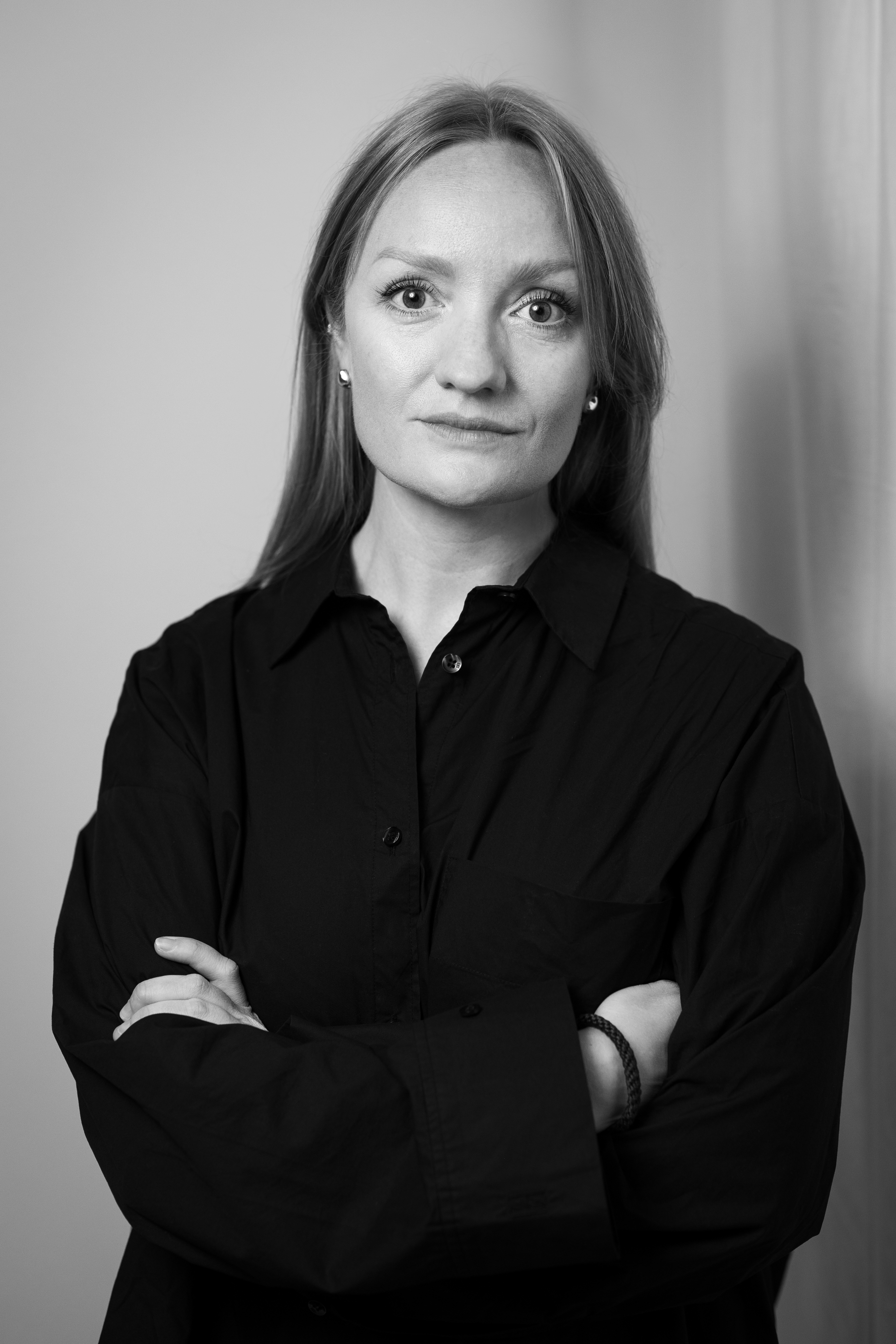 Svartvitt porträtt av en kvinna med rakt ljust hår i mörk skjorta, står med armarna i kors och tittar mot kameran.