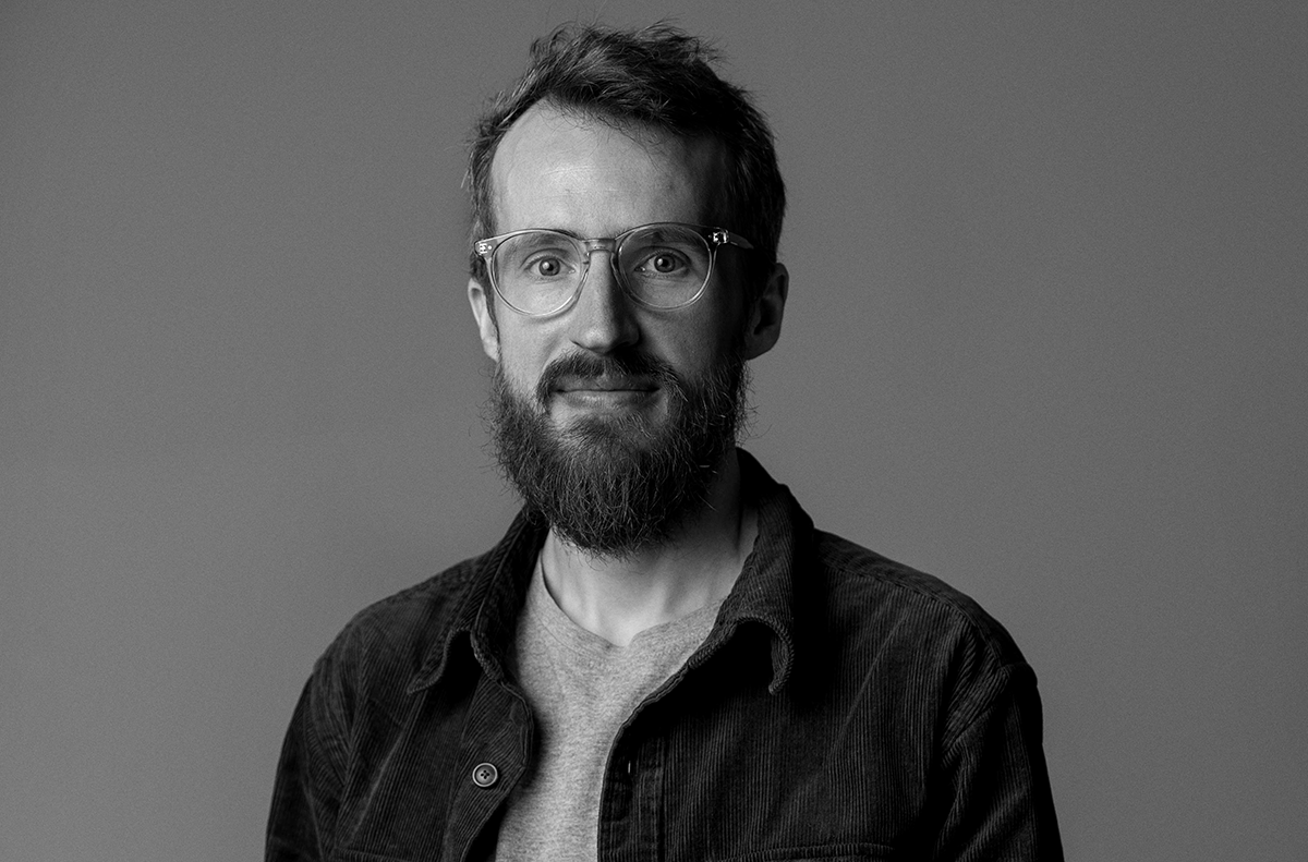 En man med glasögon och skägg har en ljus skjorta och mörk jacka och står mot en enkel bakgrund.