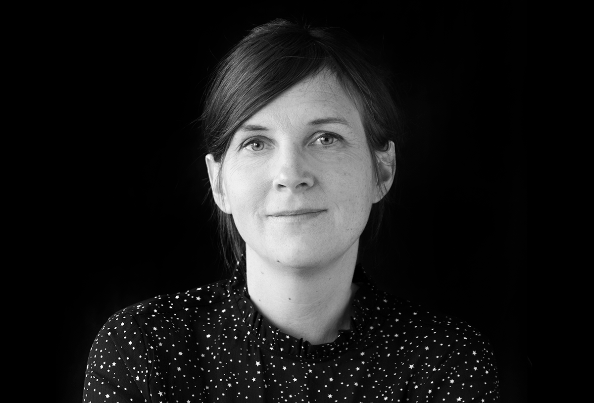 En kvinna med rakt brunt hår och en mörk, högkragad skjorta med vitt prickigt mönster ser rakt in i kameran mot svart bakgrund.