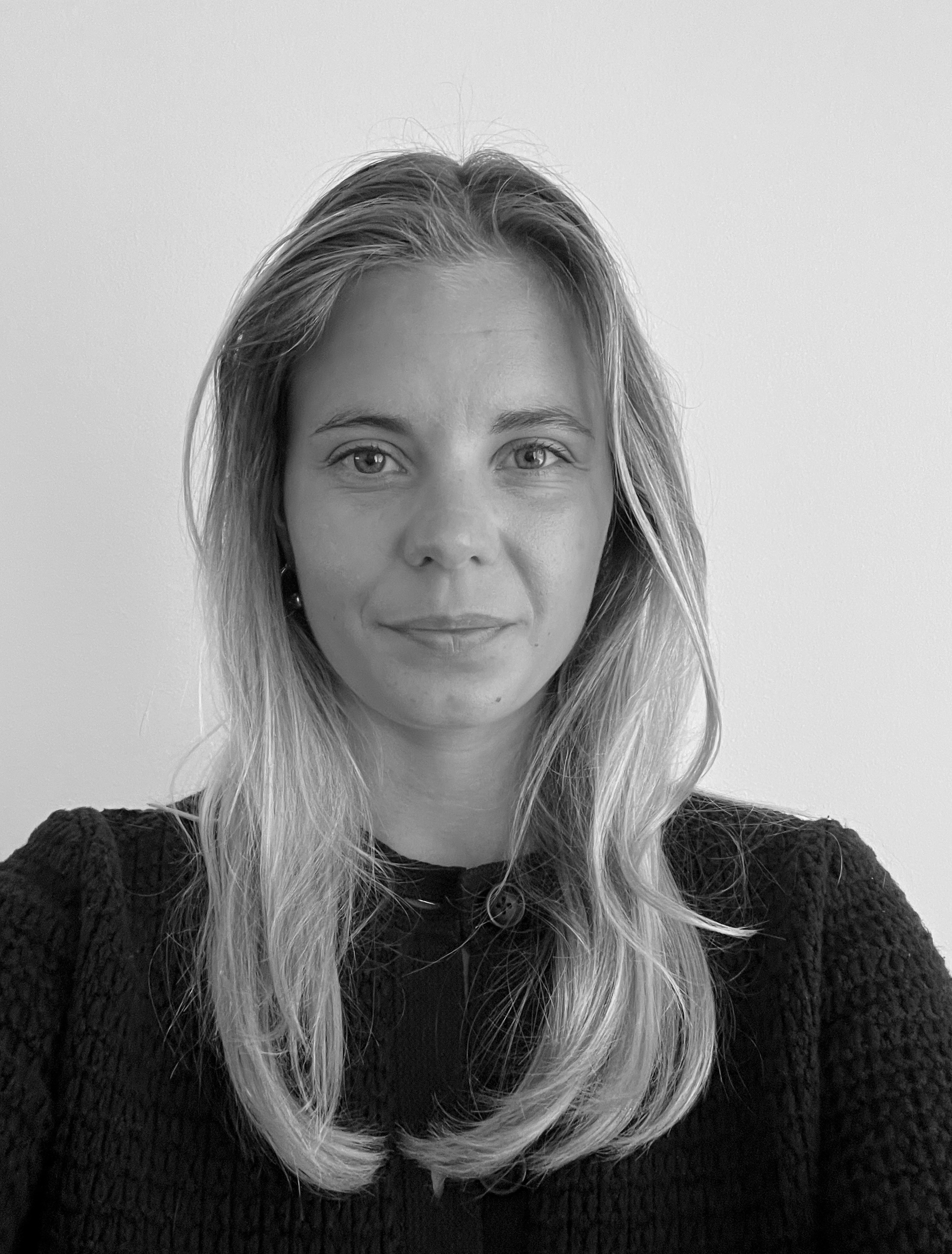 En kvinna med långt ljust hår och mörk topp ser rakt in i kameran mot en ljus bakgrund. Bilden är i svartvitt.