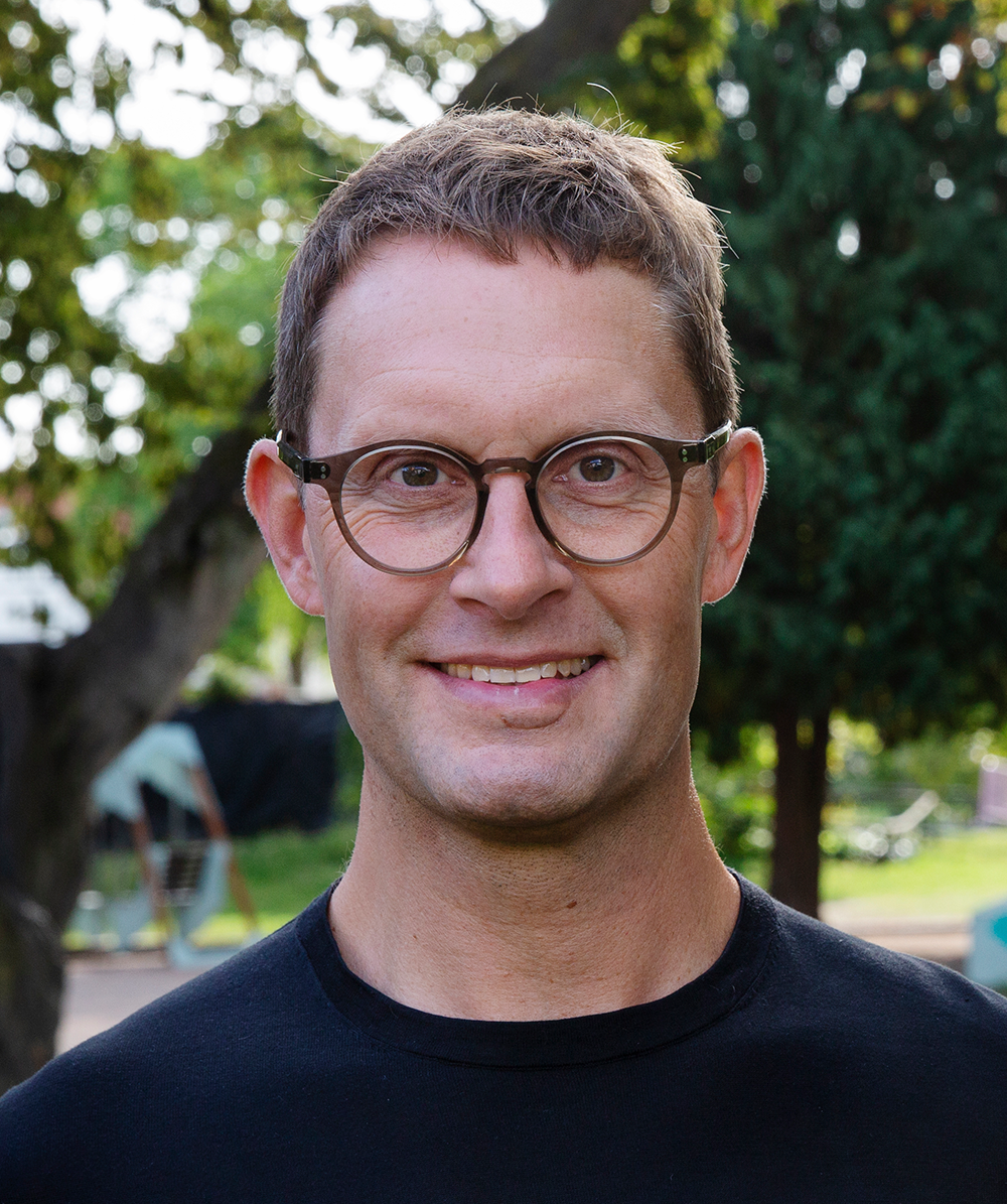 En man med kort brunt hår och glasögon har på sig en svart tröja och står utomhus med träd i bakgrunden.