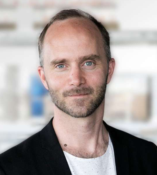 En man med kort hår och skägg, iklädd svart kavaj över vit skjorta, tittar mot kameran med neutral min. Bakgrunden är suddig.