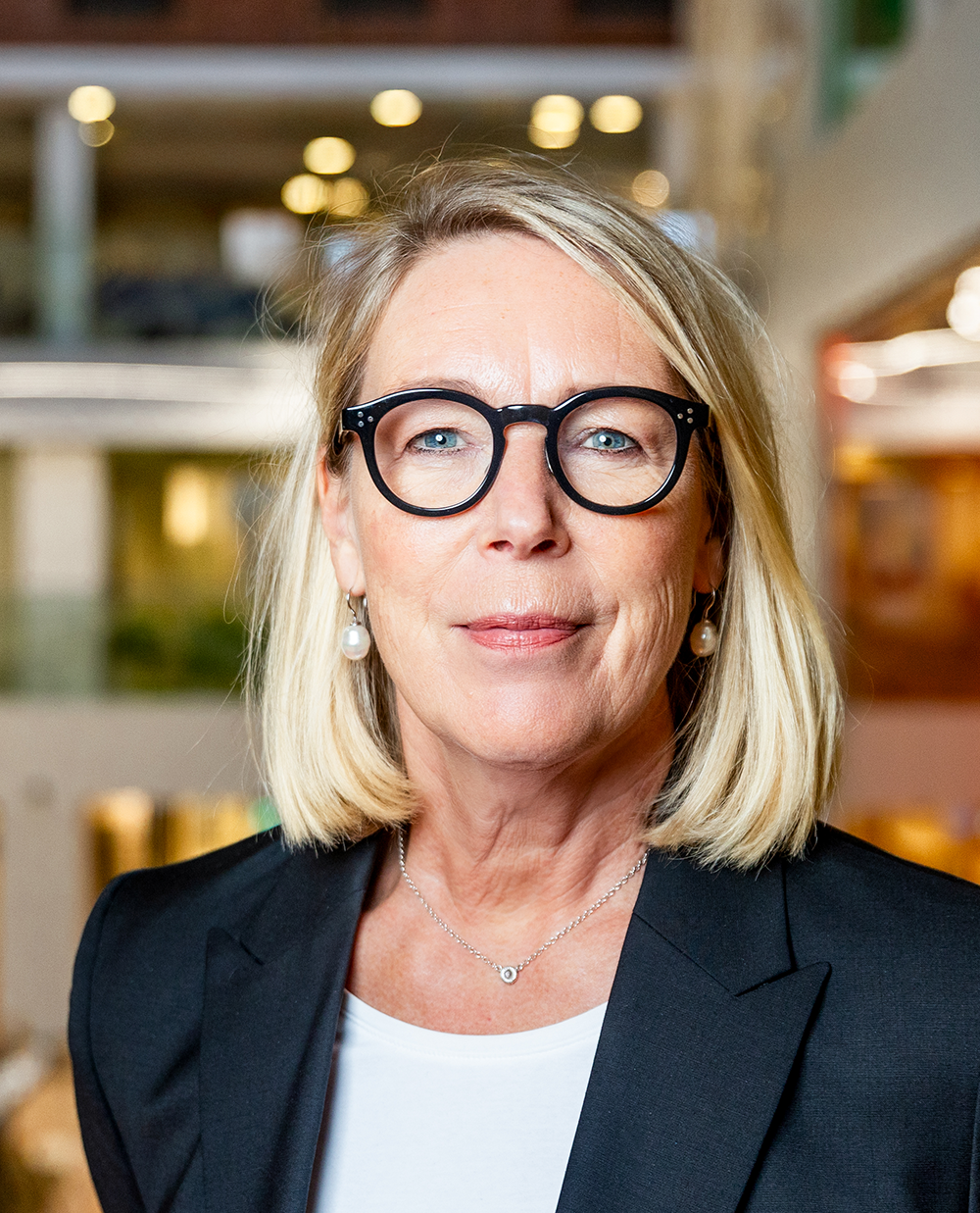 En medelålders kvinna med blont hår, glasögon och svart kavaj står inomhus i en modern miljö.