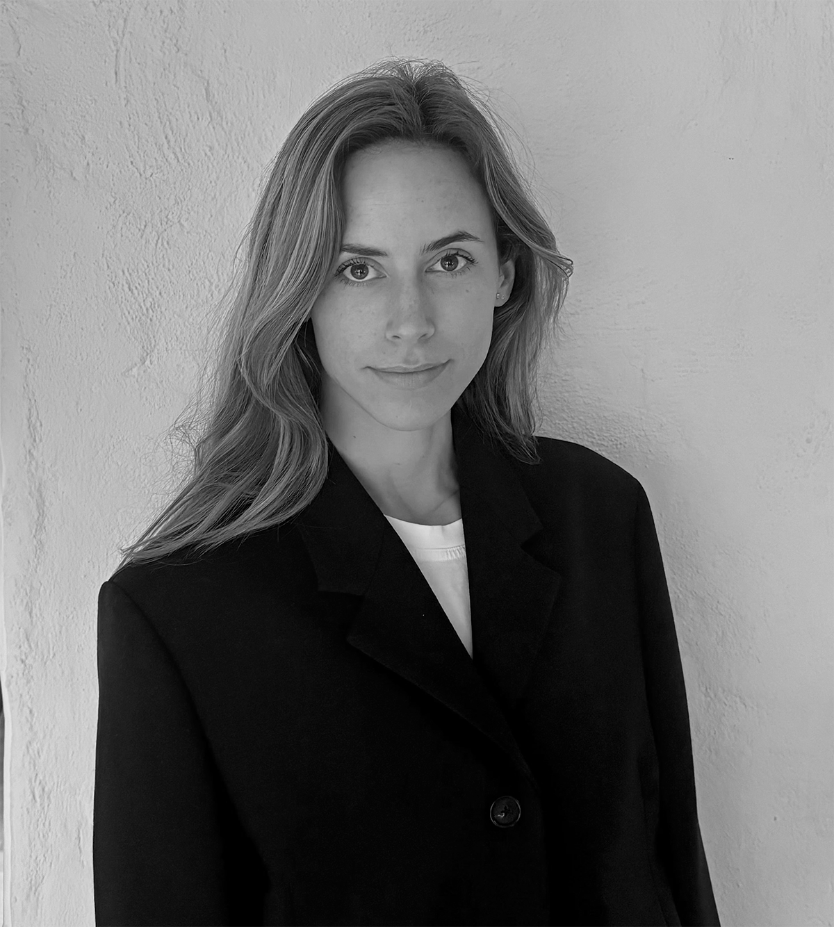 En kvinna med långt hår i mörk kavaj och vit skjorta står framför en ljus vägg och tittar mot kameran med neutral min.