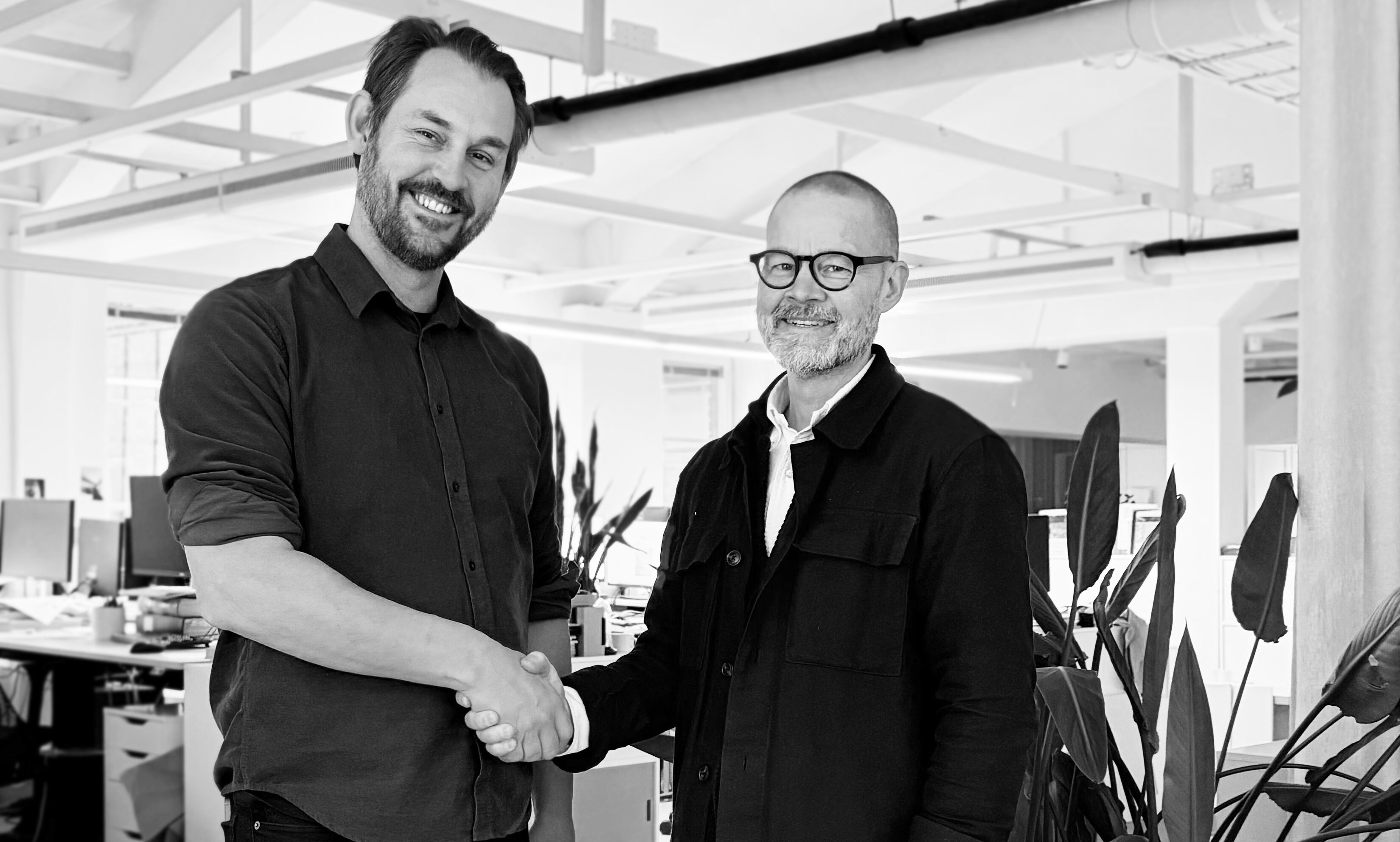 Två män skakar hand och ler i ett kontor med skrivbord, datorer och växter i bakgrunden.