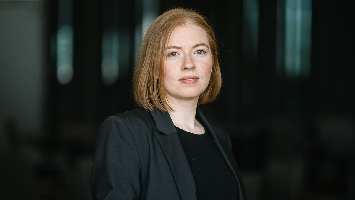 En person med rakt, ljusbrunt hår och ljus hud bär en mörk kavaj och svart topp, tittar mot kameran mot mörk bakgrund.