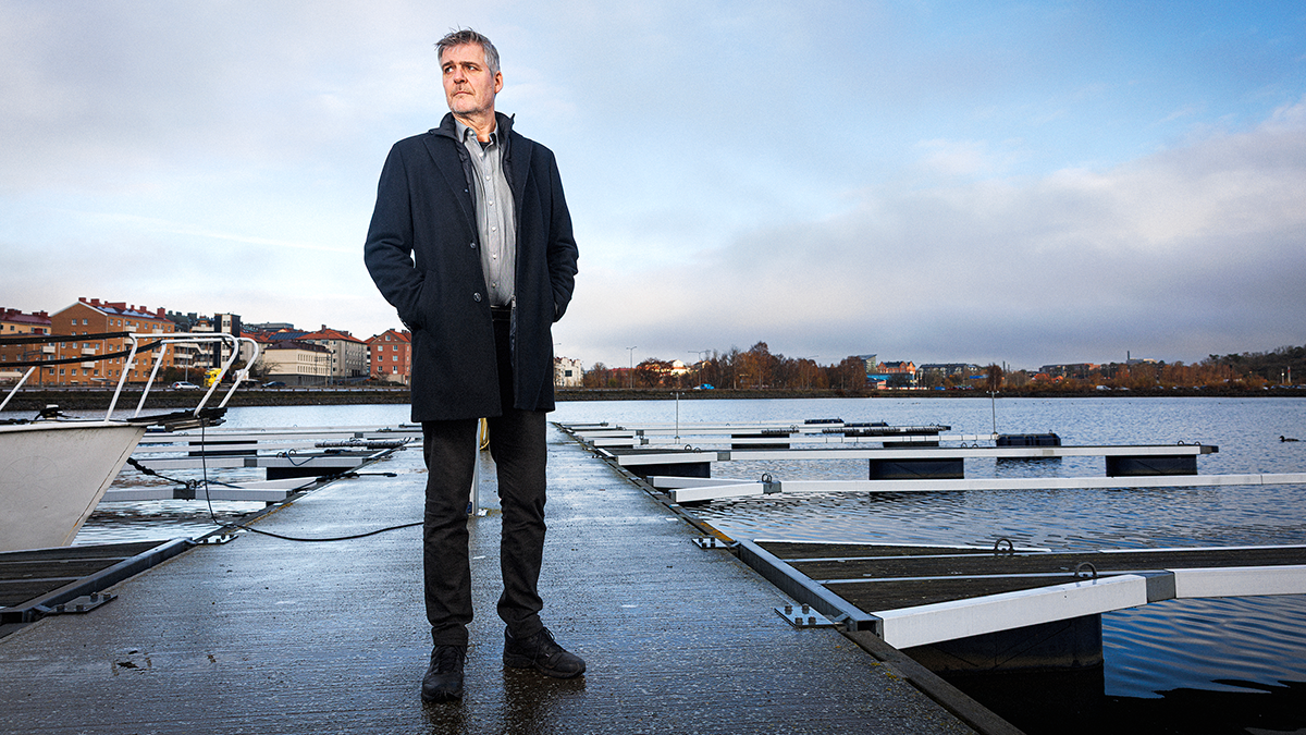 En man i en mörk kappa står på en brygga vid vattnet. I bakgrunden finns båtar och byggnader under en molnig himmel.