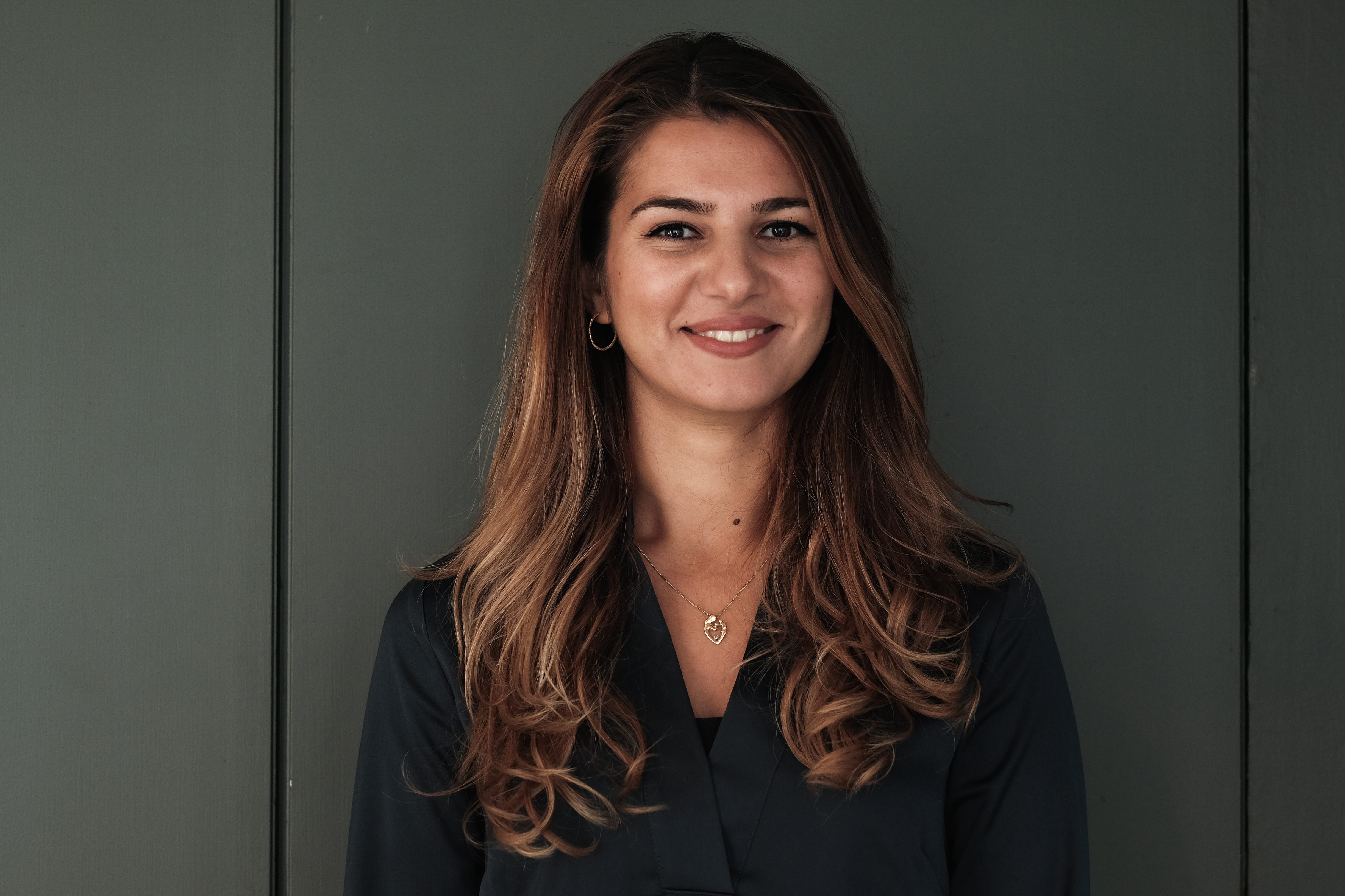 En kvinna med långt brunt hår, mörk blus och halsband står framför en mörkgrön vägg och ler mot kameran.