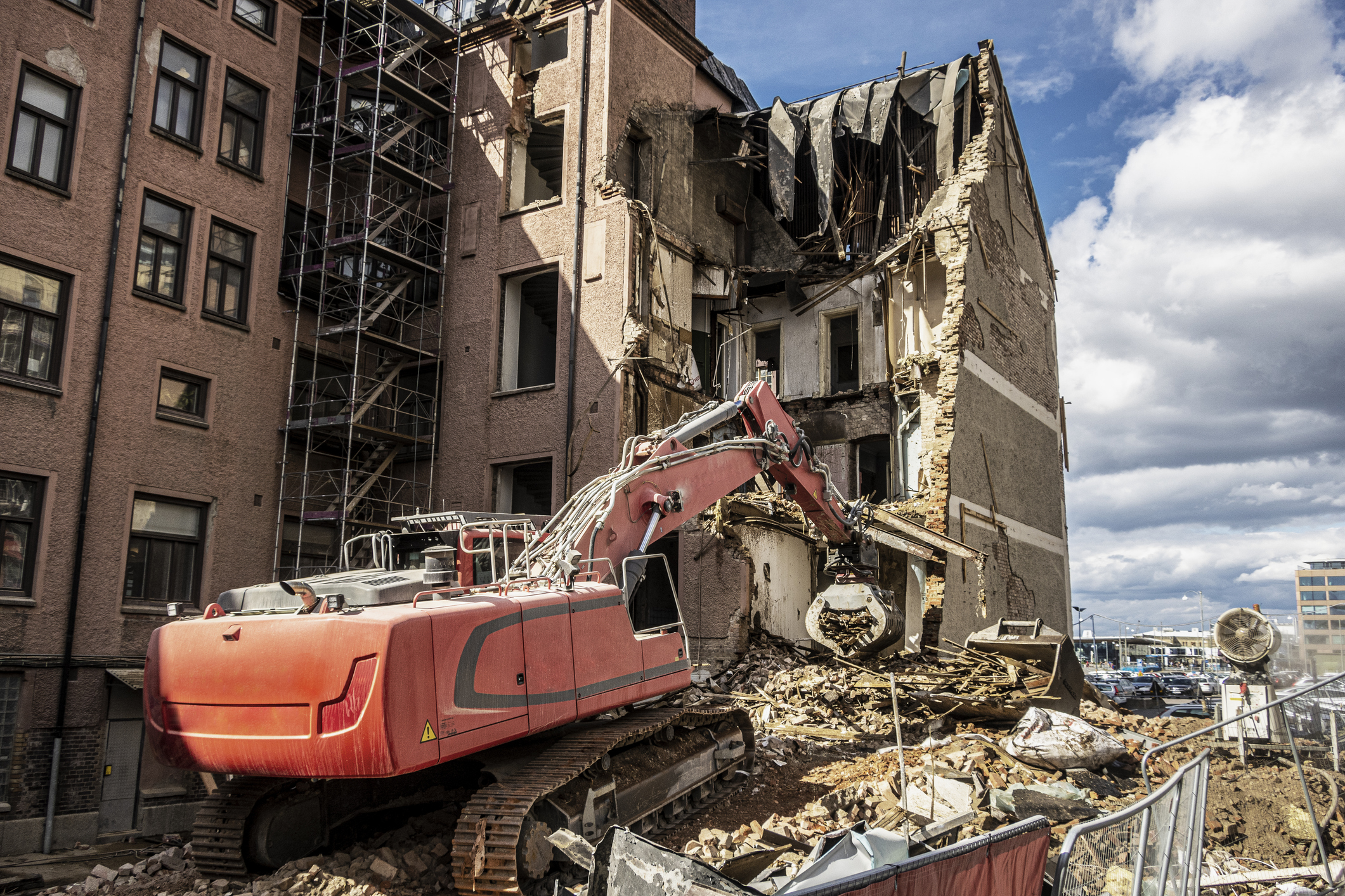 En röd grävmaskin river en del av en flervånings tegelbyggnad, med bråte utspridd kring platsen under en molnig dag.