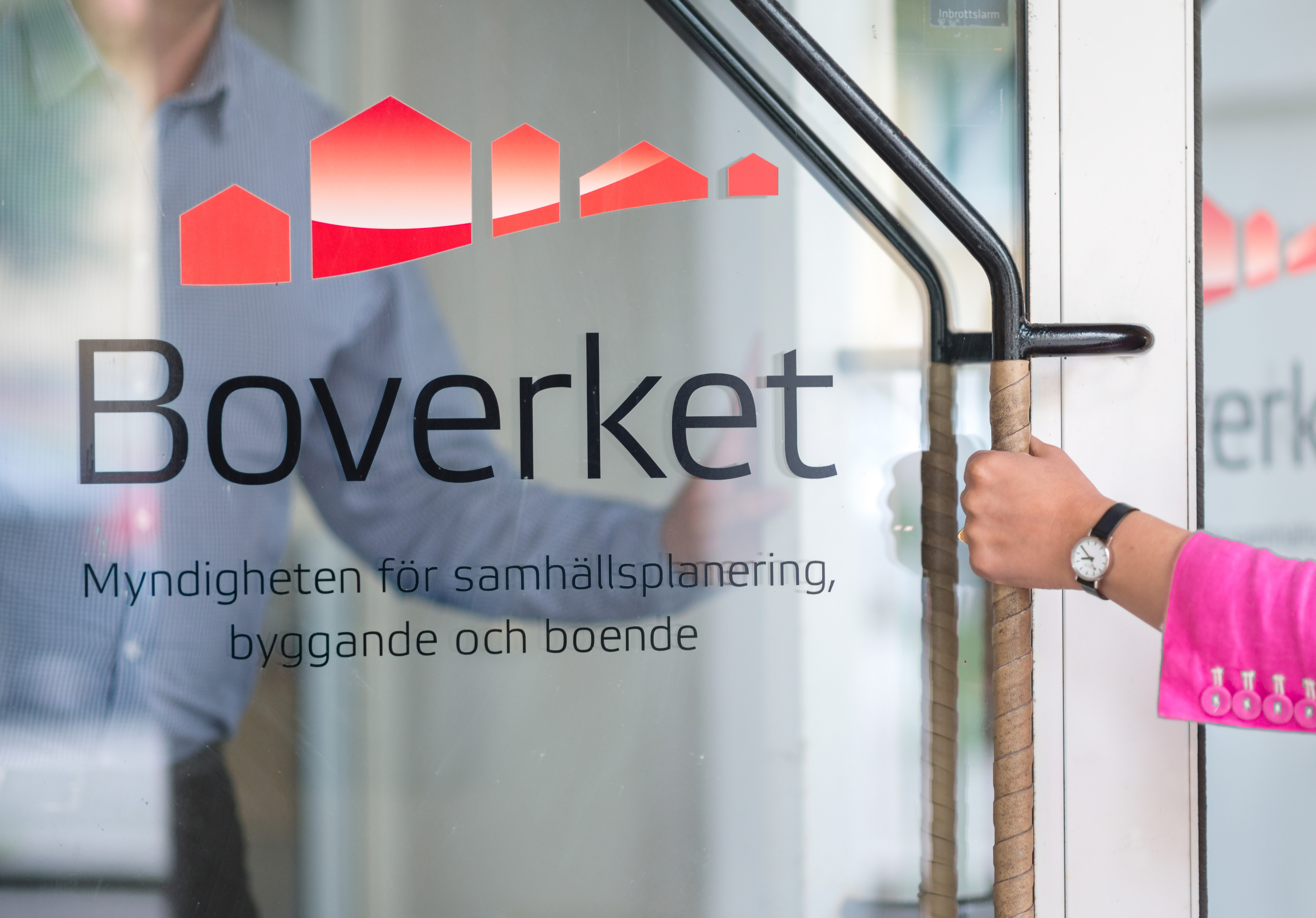 En person med rosa ärm öppnar en glasdörr med Boverkets logotyp och svensk text om samhällsplanering, byggande och boende.