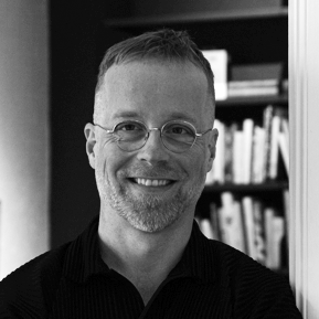 En man med glasögon och kort skägg ler mot kameran; bokhyllor syns i bakgrunden.