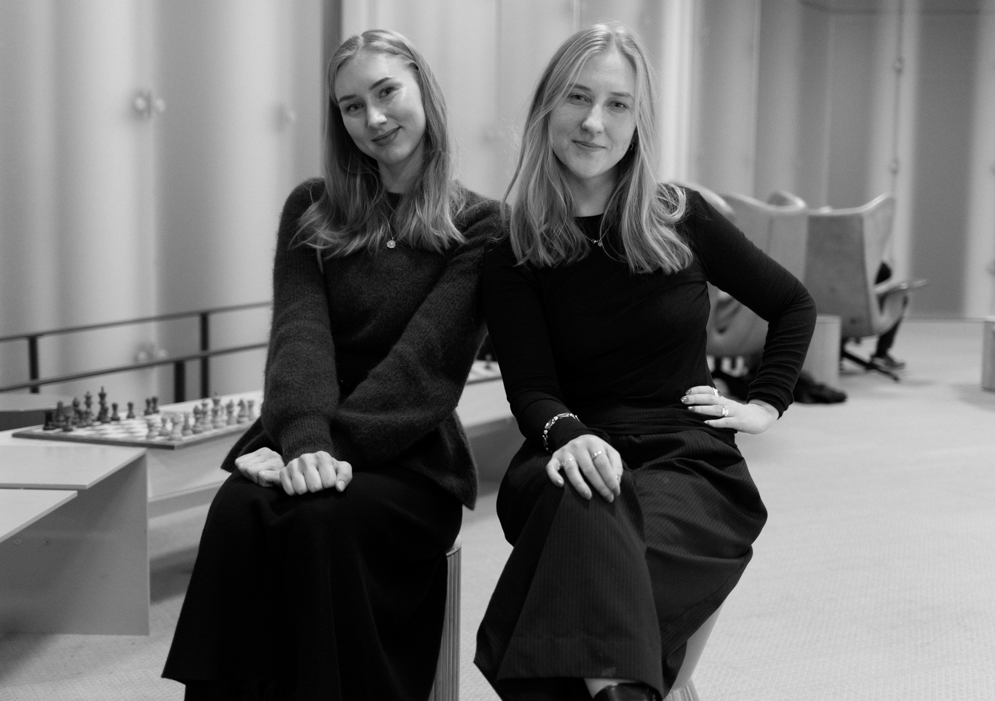 Två kvinnor sitter bredvid varandra inomhus, vända mot kameran och ler svagt. Ett schackbräde syns på bordet bakom dem. Bilden är i svartvitt.