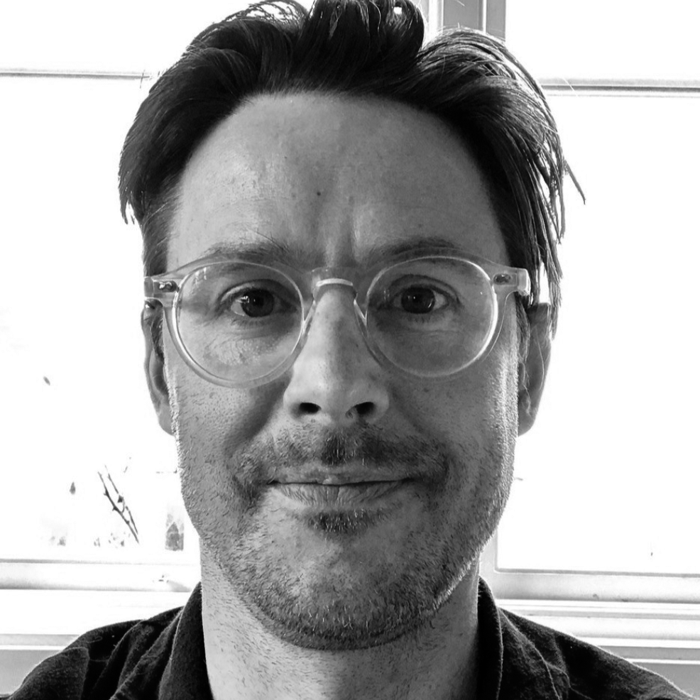 En man med kort mörkt hår, glasögon och lätt skägg sitter inomhus framför ett fönster och tittar mot kameran. Svartvitt foto.