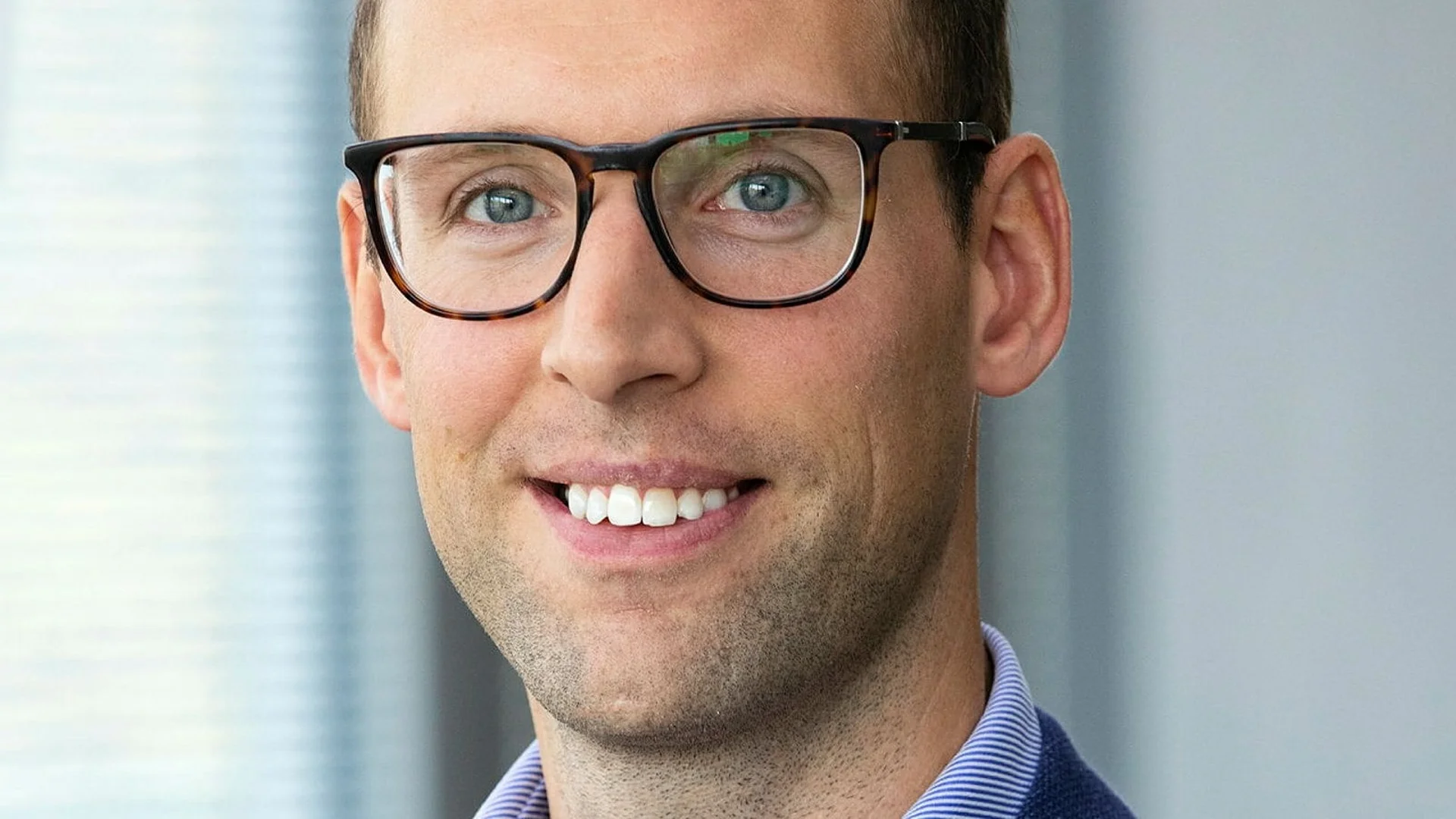 En man med kort brunt hår, har glasögon och en blå skjorta med krage, tittar mot kameran inomhus.