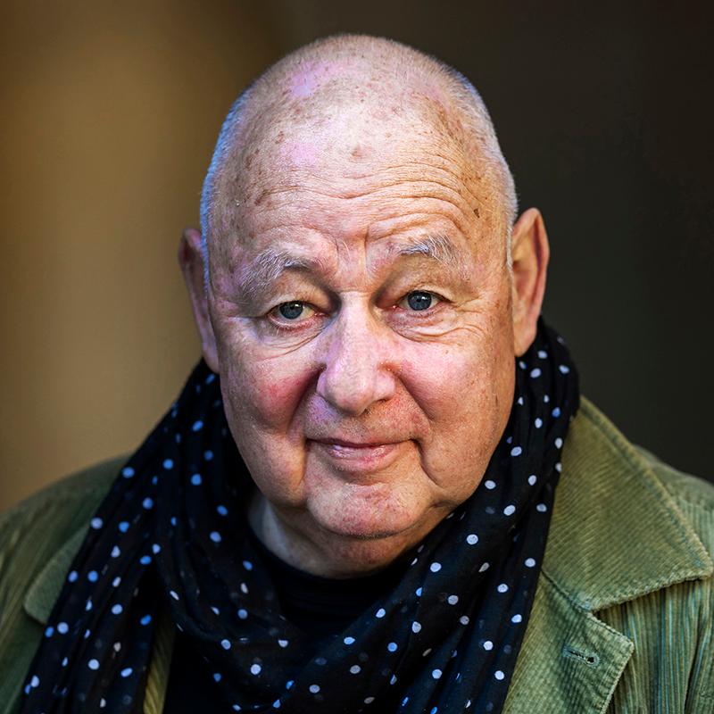 En äldre man med rakat huvud har en grön jacka och en svart scarf med vita prickar. Han tittar in i kameran och ler lite.