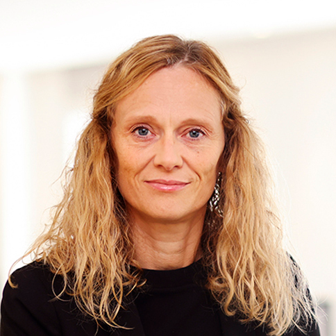 En kvinna med vågigt blont hår och svart topp tittar rakt mot kameran mot en neutralt belyst bakgrund.