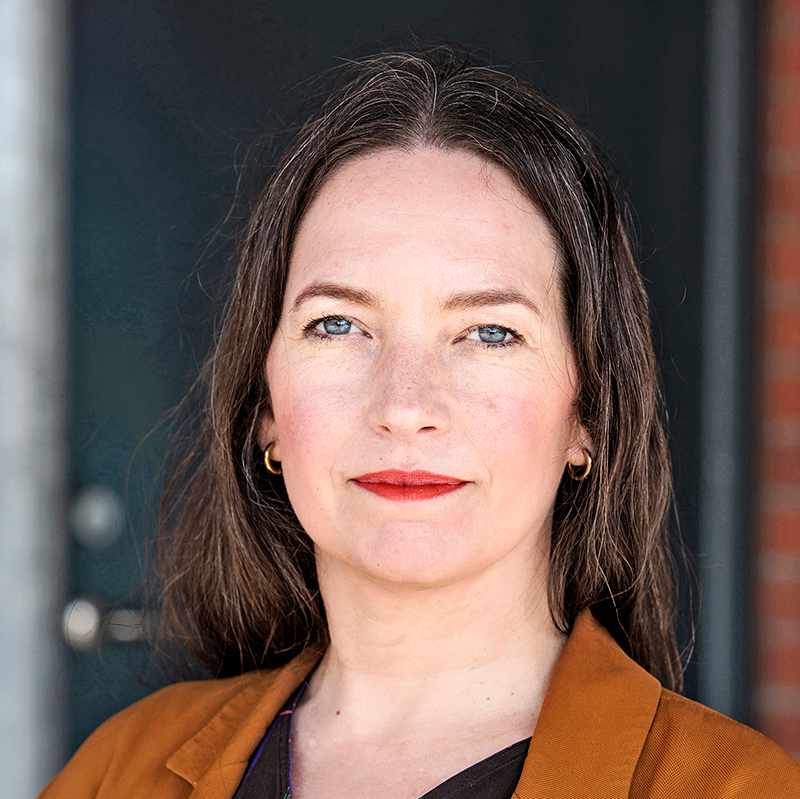 En kvinna med långt brunt hår och rött läppstift ser in i kameran. Hon har en brun kavaj och små ringörhängen.