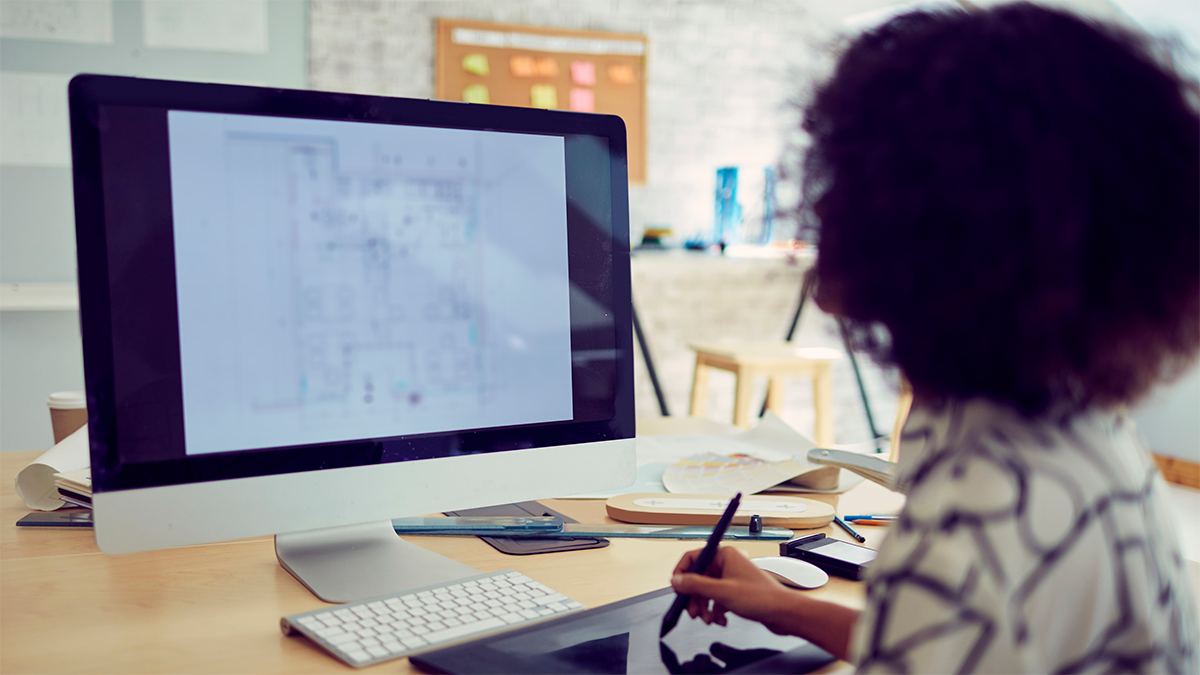 En person sitter vid ett skrivbord och använder en digital penna samt ritplatta medan arkitektritningar visas på en datorskärm.