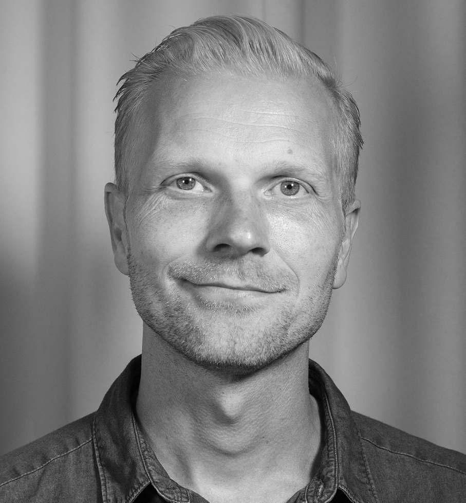 En man med kort ljust hår och välansad skäggväxt ser mot kameran och ler svagt. Han har på sig en skjorta. Bakgrunden är enkel.