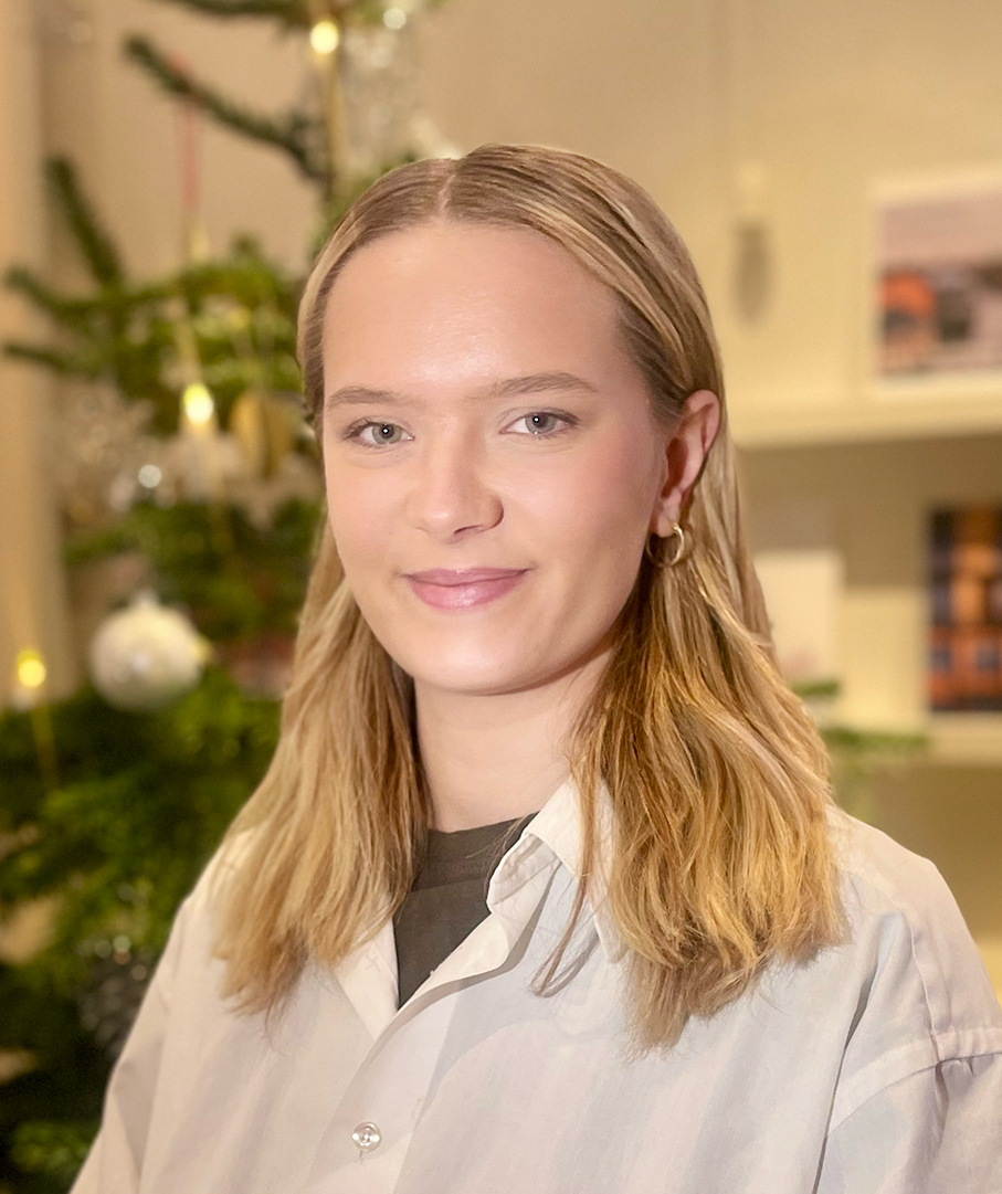 En kvinna med rakt blont hår och vit skjorta står inomhus framför en julgran med dekorationer.