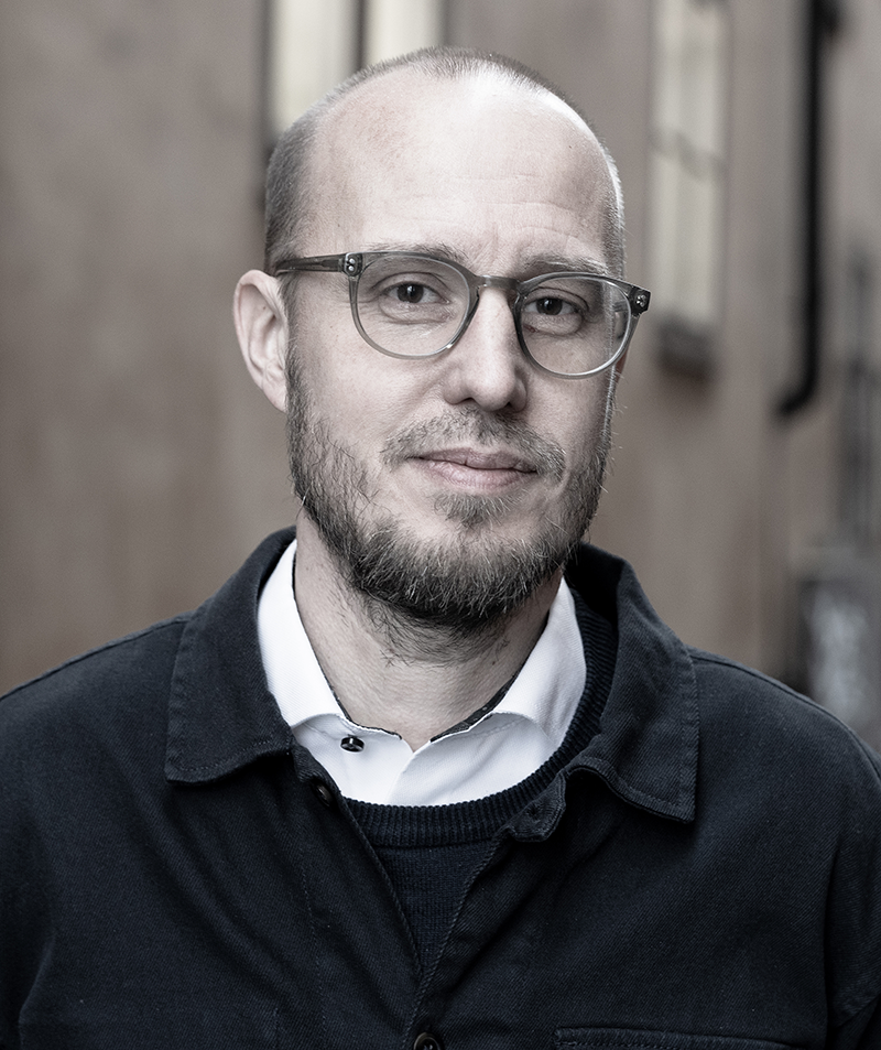 En man med glasögon och skägg står utomhus med neutral min. Han har vit skjorta och mörk jacka. Bakgrunden är suddig.