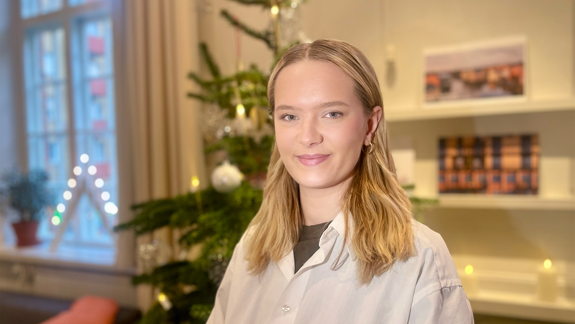 En kvinna med blont hår och vit skjorta står inomhus framför en julgran, med hyllor och ett fönster i bakgrunden.