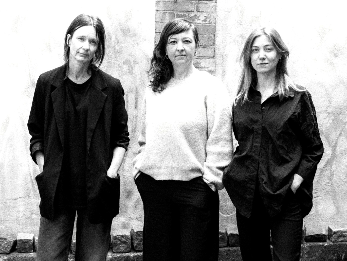 Tre kvinnor står bredvid varandra framför en vägg, vända mot kameran med händerna i fickorna och neutrala ansiktsuttryck.