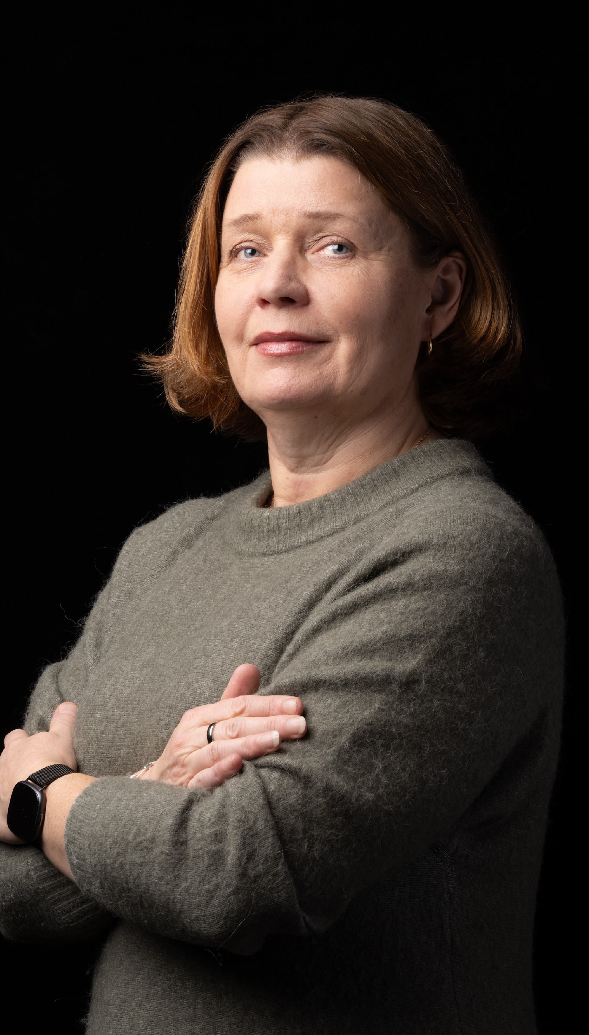 Kvinna i medelåldern med kort brunt hår, grå tröja och smartklocka står med korslagda armar mot svart bakgrund.