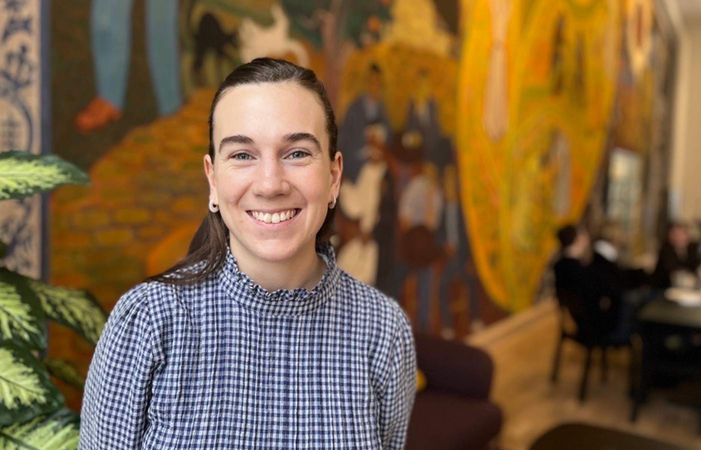 En kvinna i en rutig skjorta ler mot kameran, står inomhus med en väggmålning och flera personer i bakgrunden.