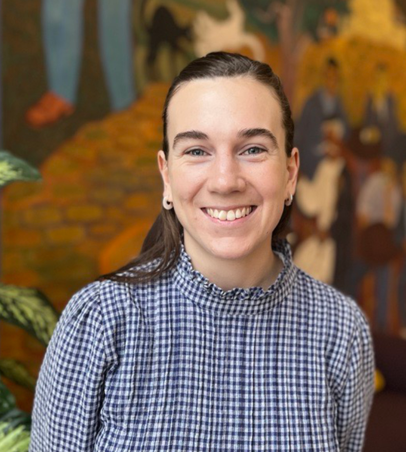 En kvinna med brunt hår i en rutig skjorta ler mot kameran, står inomhus med en muralmålning i bakgrunden.