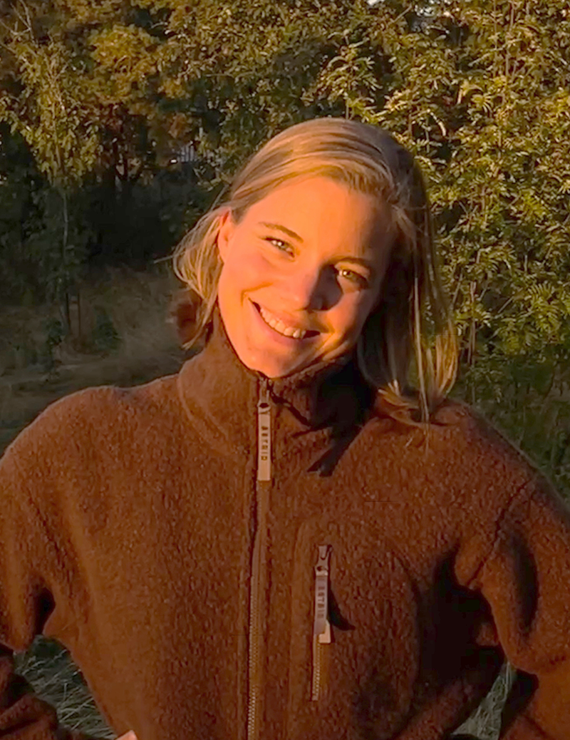 En person med en brun fleecejacka står utomhus, ler, med solen på ansiktet och träd i bakgrunden.