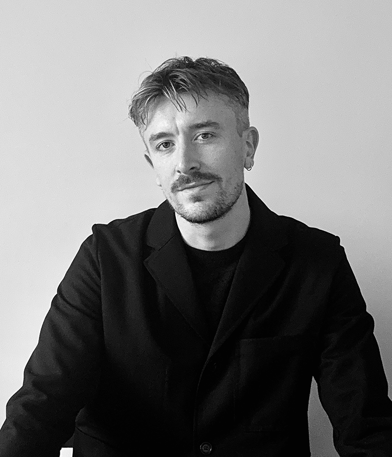 En man med kort hår och ansiktshår sitter mot en ljus bakgrund, iförd en mörk jacka, och tittar mot kameran.