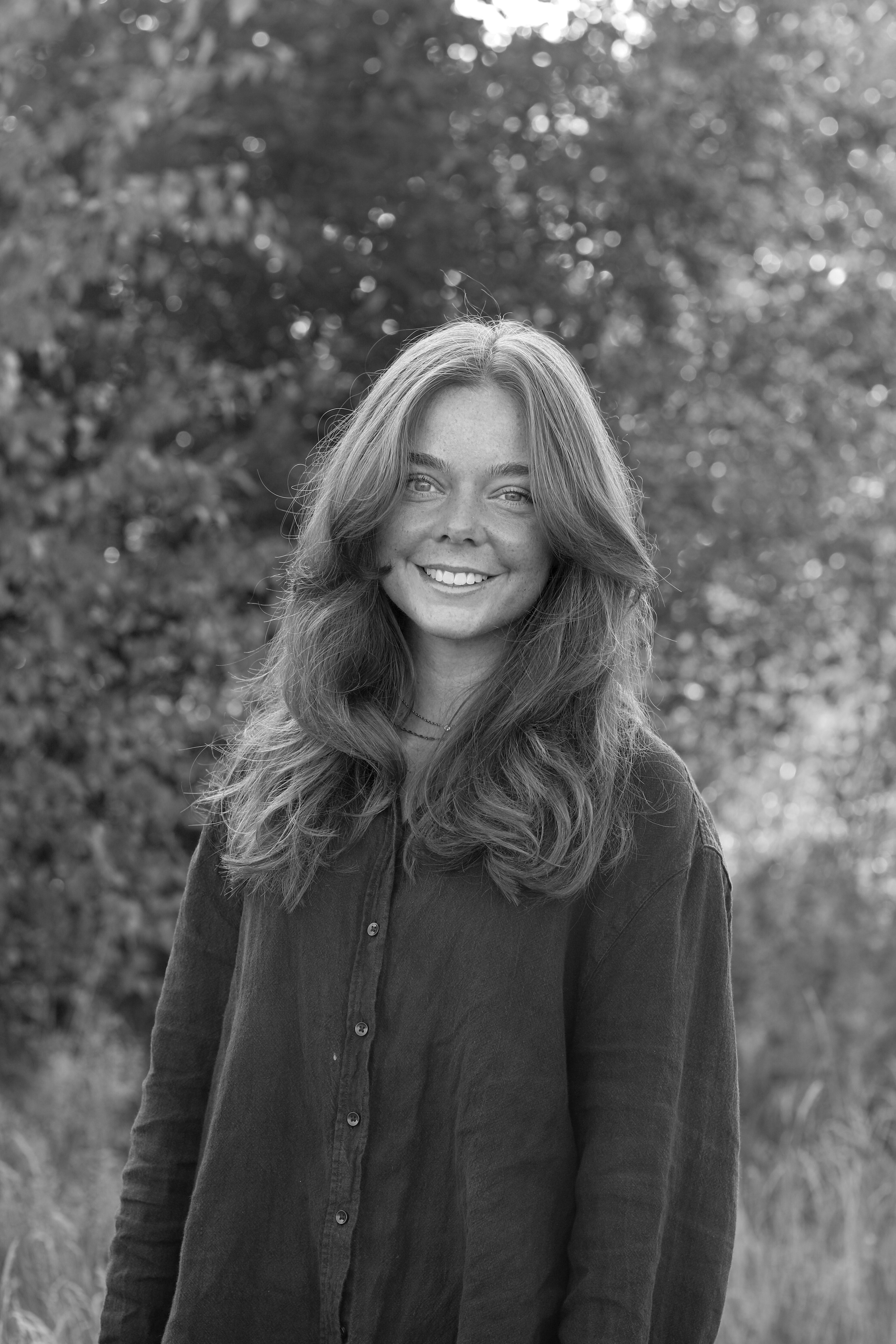 En ung kvinna med långt, vågigt hår står utomhus och ler mot kameran. Hon har en skjorta på sig. Bakomom syns lövverk. Fotot är i svartvitt.
