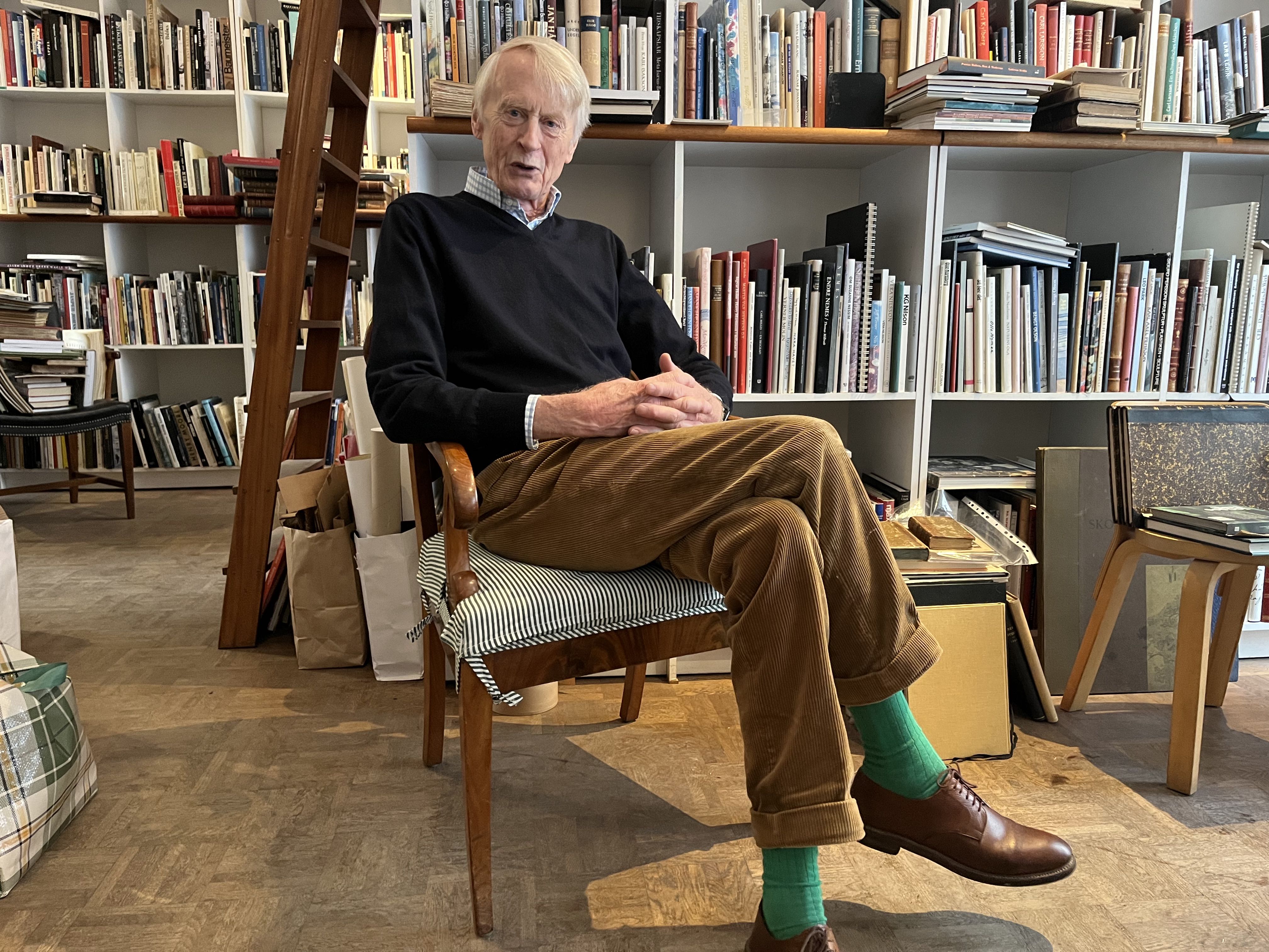 En äldre man i svart tröja, bruna byxor och gröna strumpor sitter på en stol i ett rum med bokhyllor fyllda med böcker.