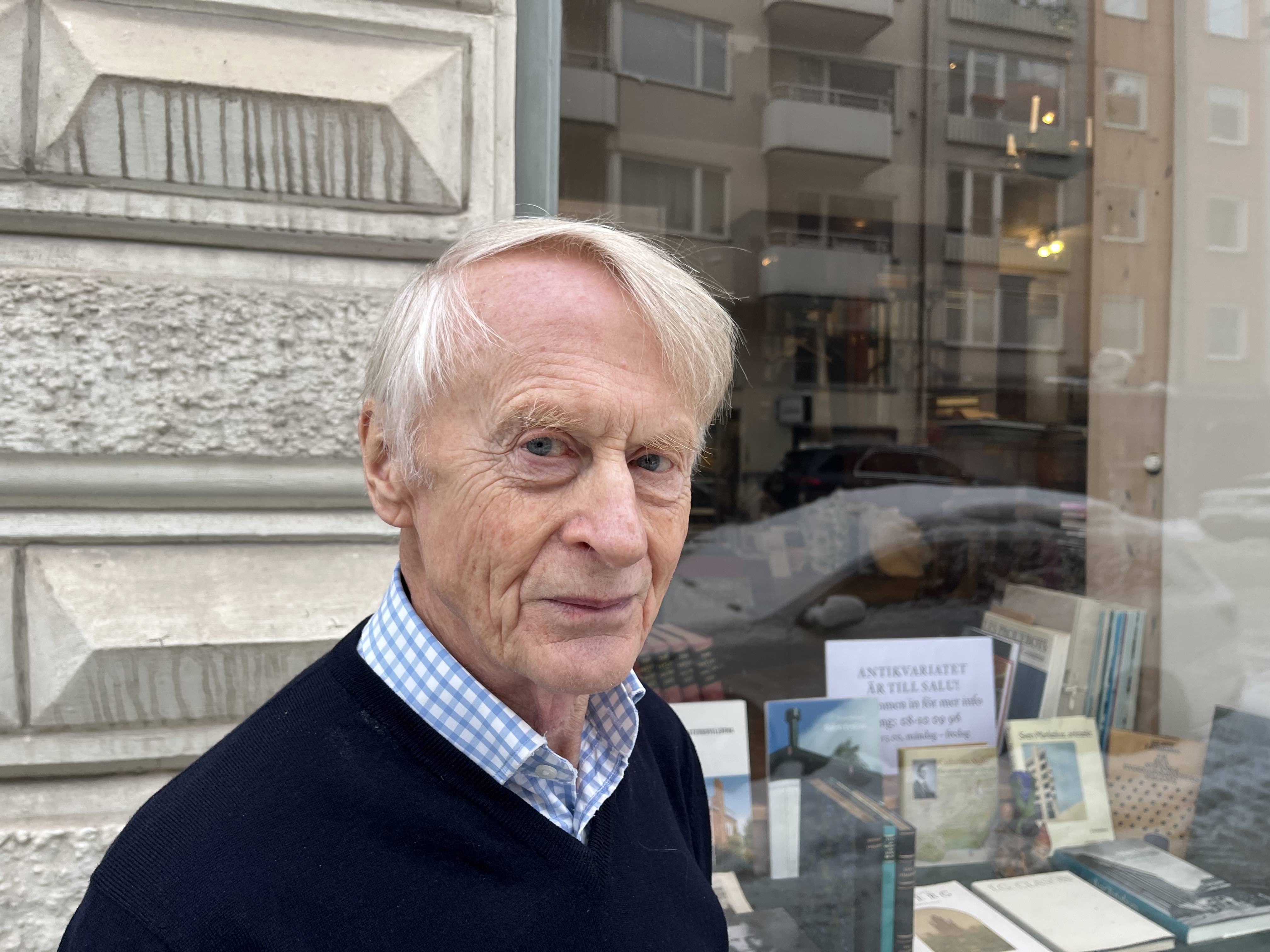 En äldre man med ljust hår står framför ett bokskyltfönster där böcker och en skylt syns, samt byggnader speglas i rutan.
