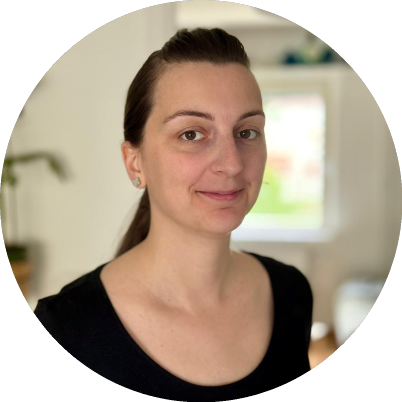 En kvinna med bakåtuppsatt mörkt hår och svart topp står inomhus framför ett fönster och tittar mot kameran.