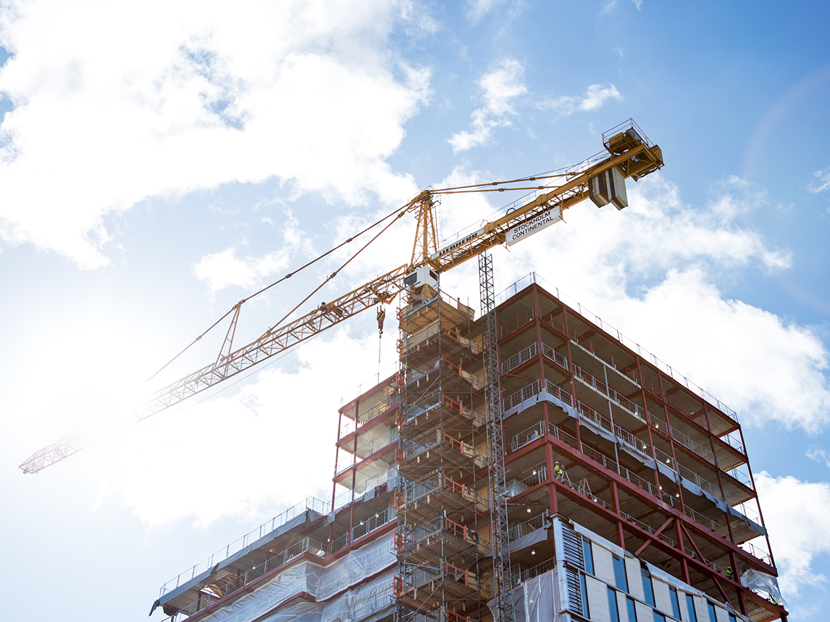 En kran står bredvid en byggnad med flera våningar av stål och betong som är under konstruktion mot en blå himmel med moln.