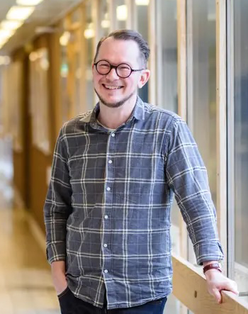 En person med glasögon och rutig skjorta står inomhus vid ett fönster, ler och har händerna i fickorna.